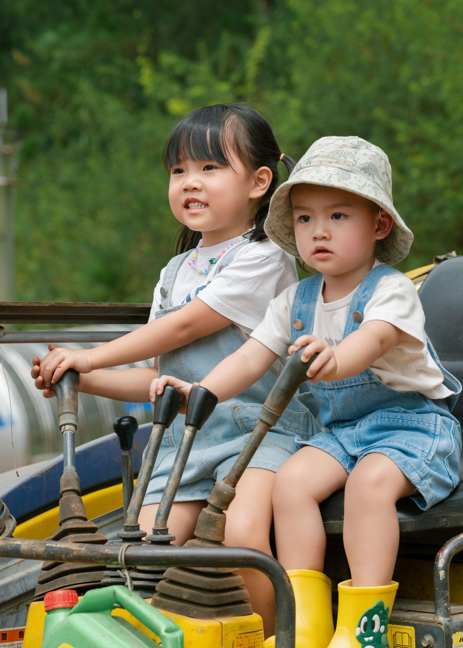 Two young Asian children in matching denim overalls sitting on a tractor, surrounded by green foliage.