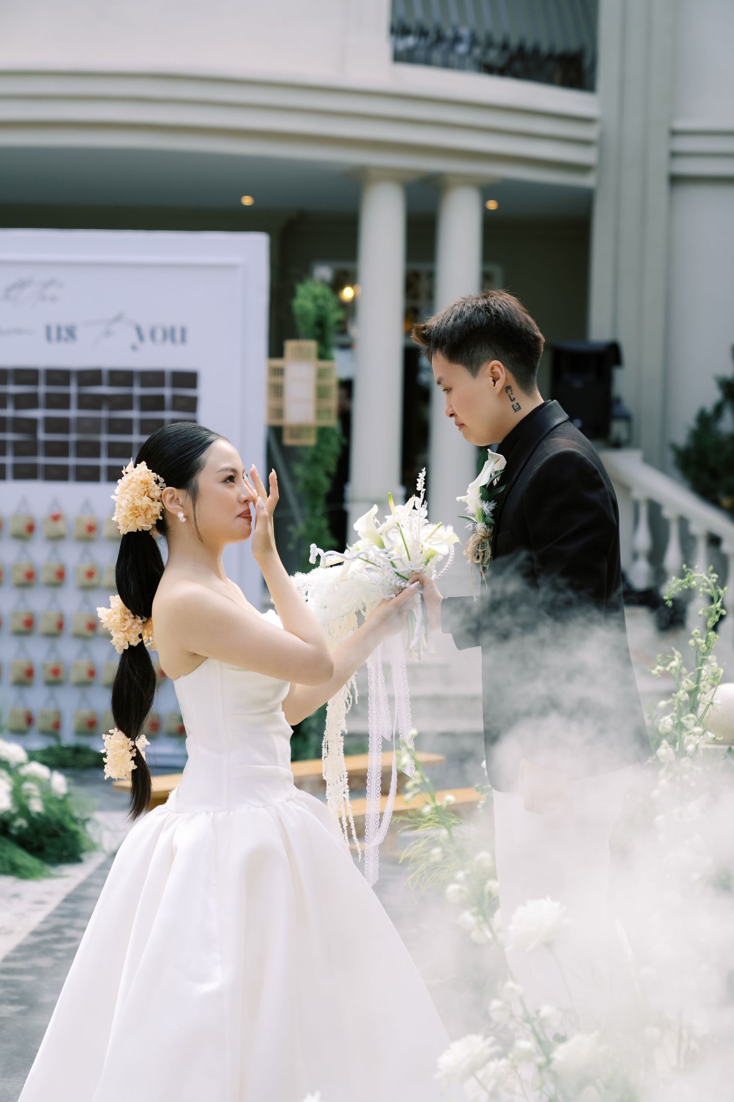 Bride in white gown holds bouquet while groom in black suit stands opposite during outdoor wedding ceremony.