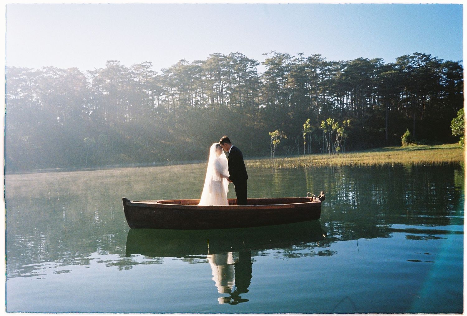 Bride and groom share a tender moment in a wooden rowboat on a calm reflective lake surrounded by pines.