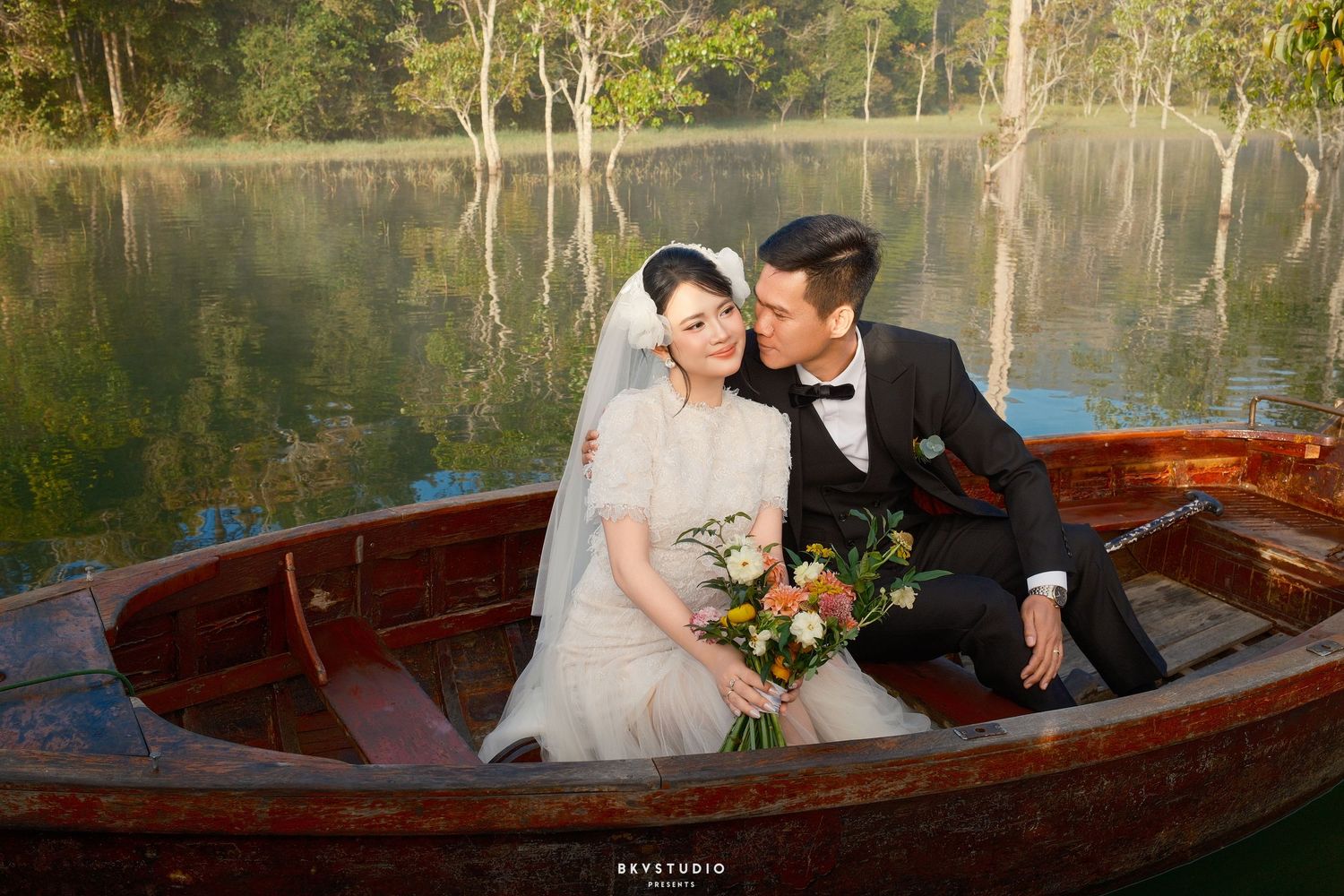 Bride and groom share a romantic moment in a wooden rowboat on a serene reflective lake.
