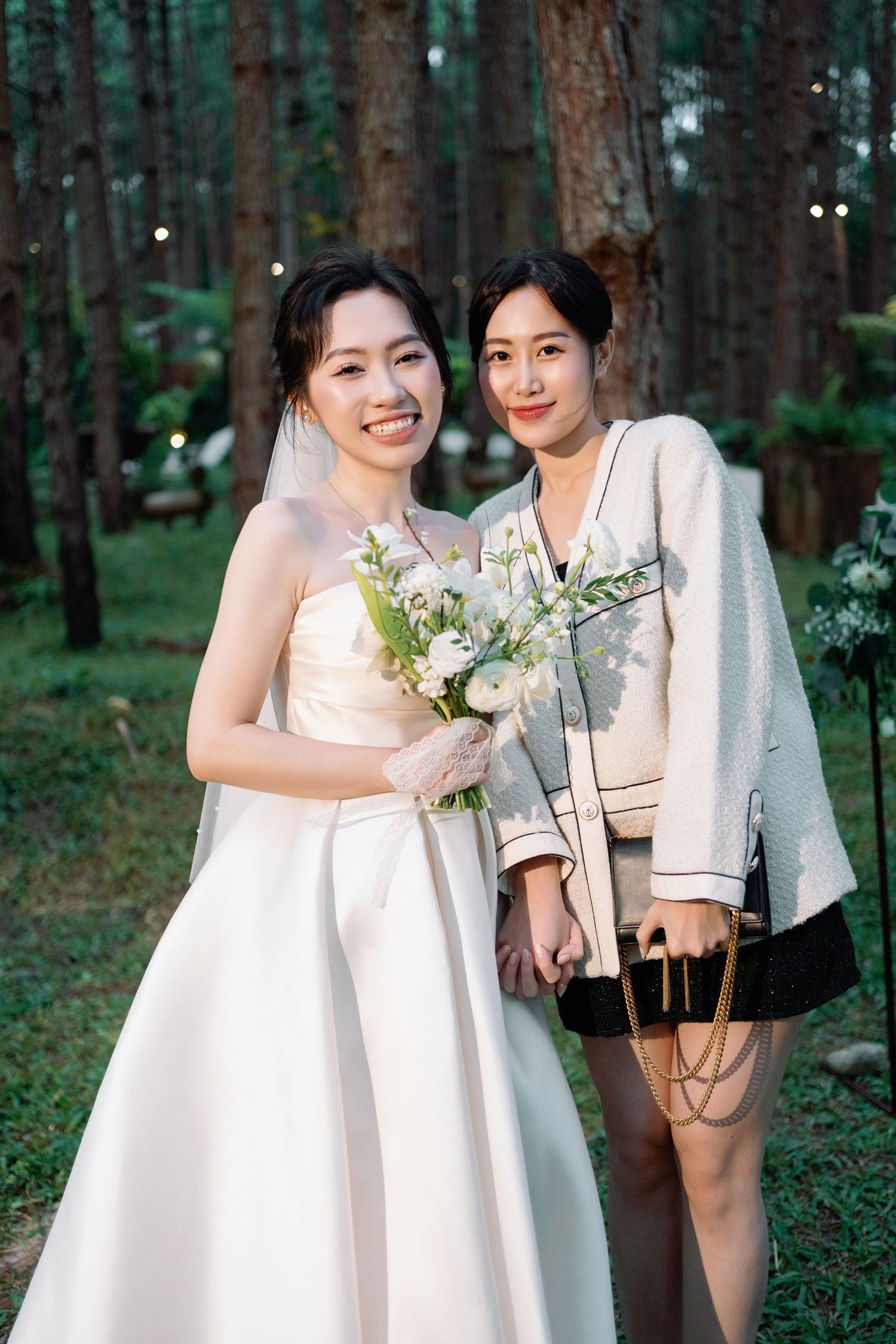 Bride in strapless white wedding gown holding bouquet poses with a female guest in a pine forest setting.