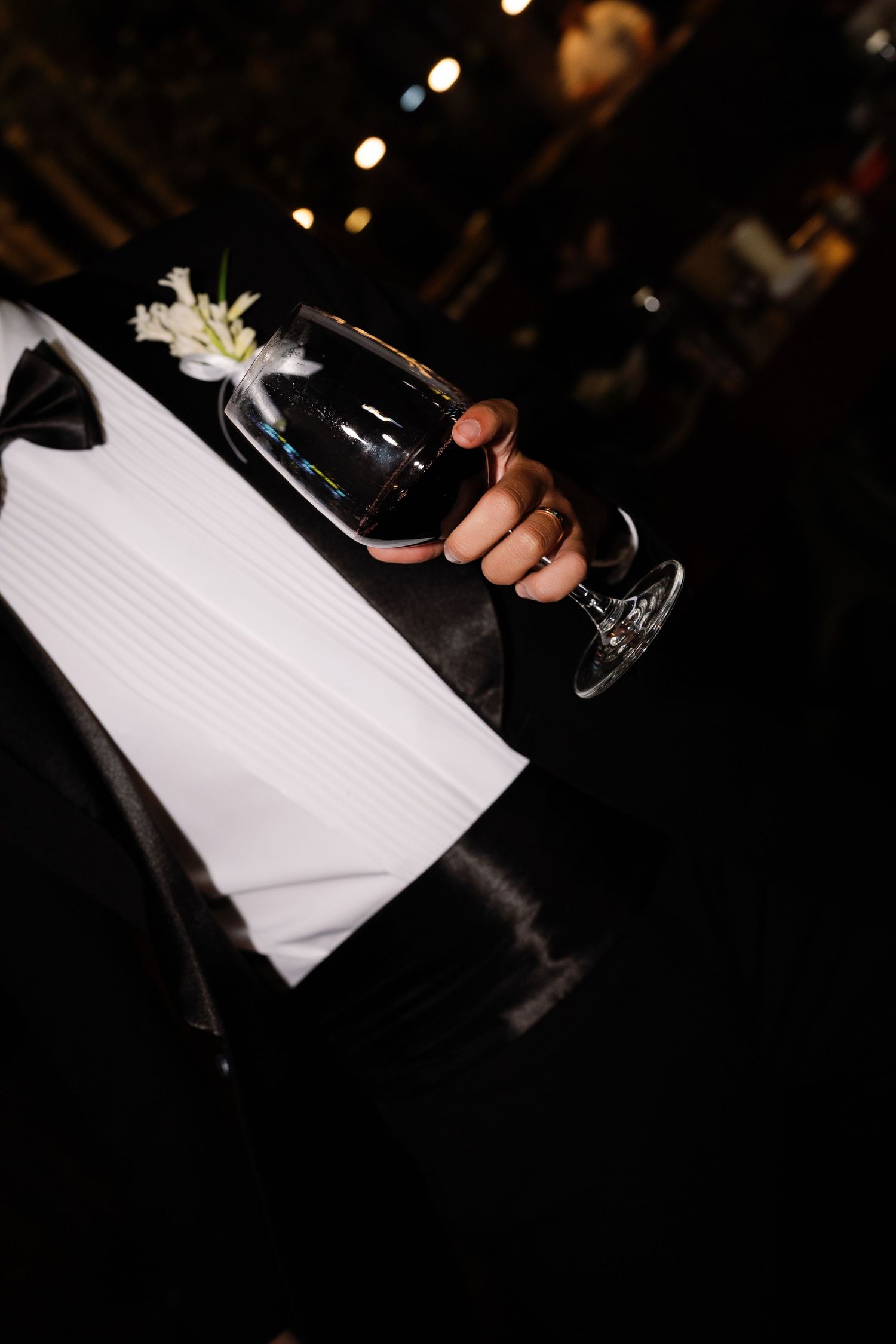 Groom in black tuxedo holding crystal wine glass at elegant evening wedding reception.