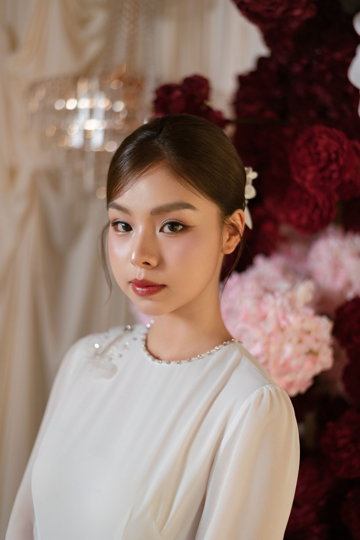 Young Asian woman in white pearl-embellished dress poses elegantly before lush red and pink floral backdrop.