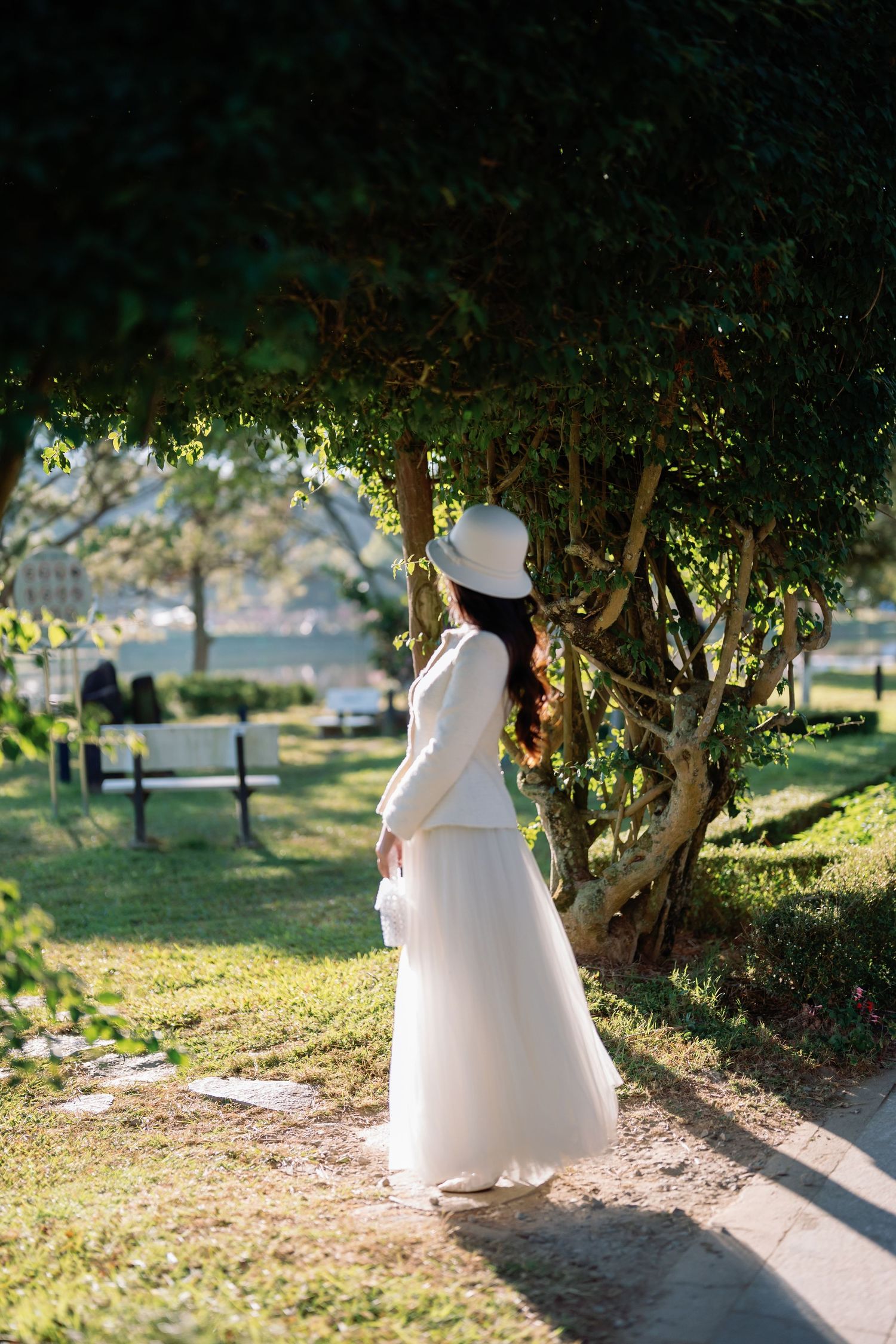 Woman in white dress and hat standing by lush green tree in sunlit park garden.