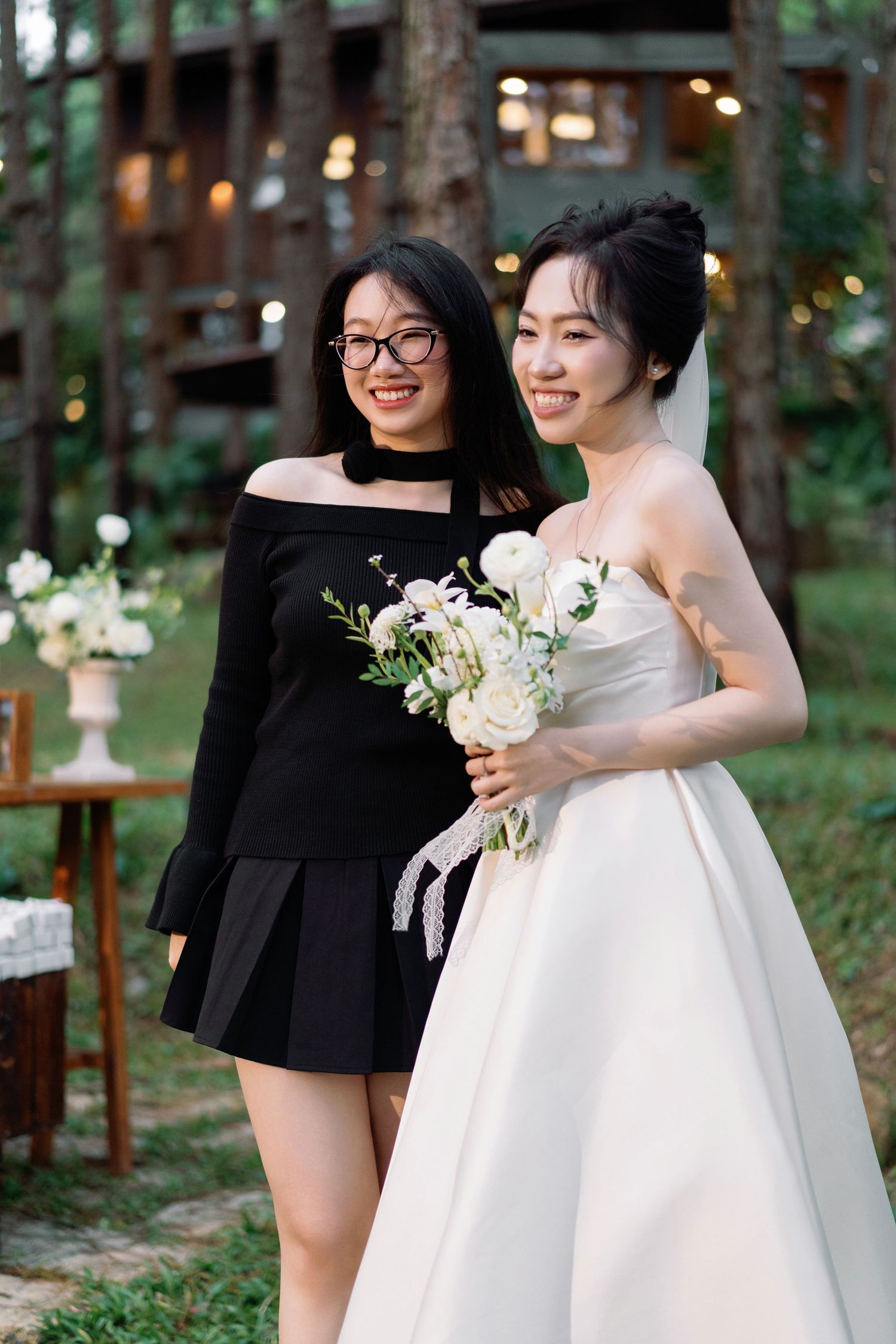Bride in white strapless gown holding white bouquet posing with a guest in black dress at outdoor wedding.