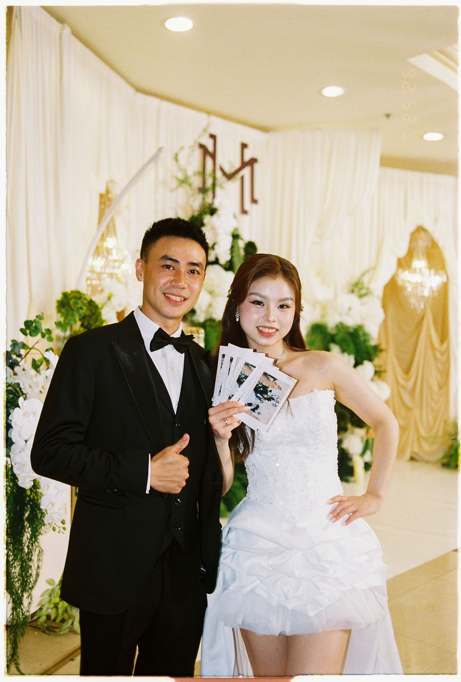 Couple posing at elegant wedding venue, man in tuxedo giving thumbs up, woman in white dress holding photo strips.