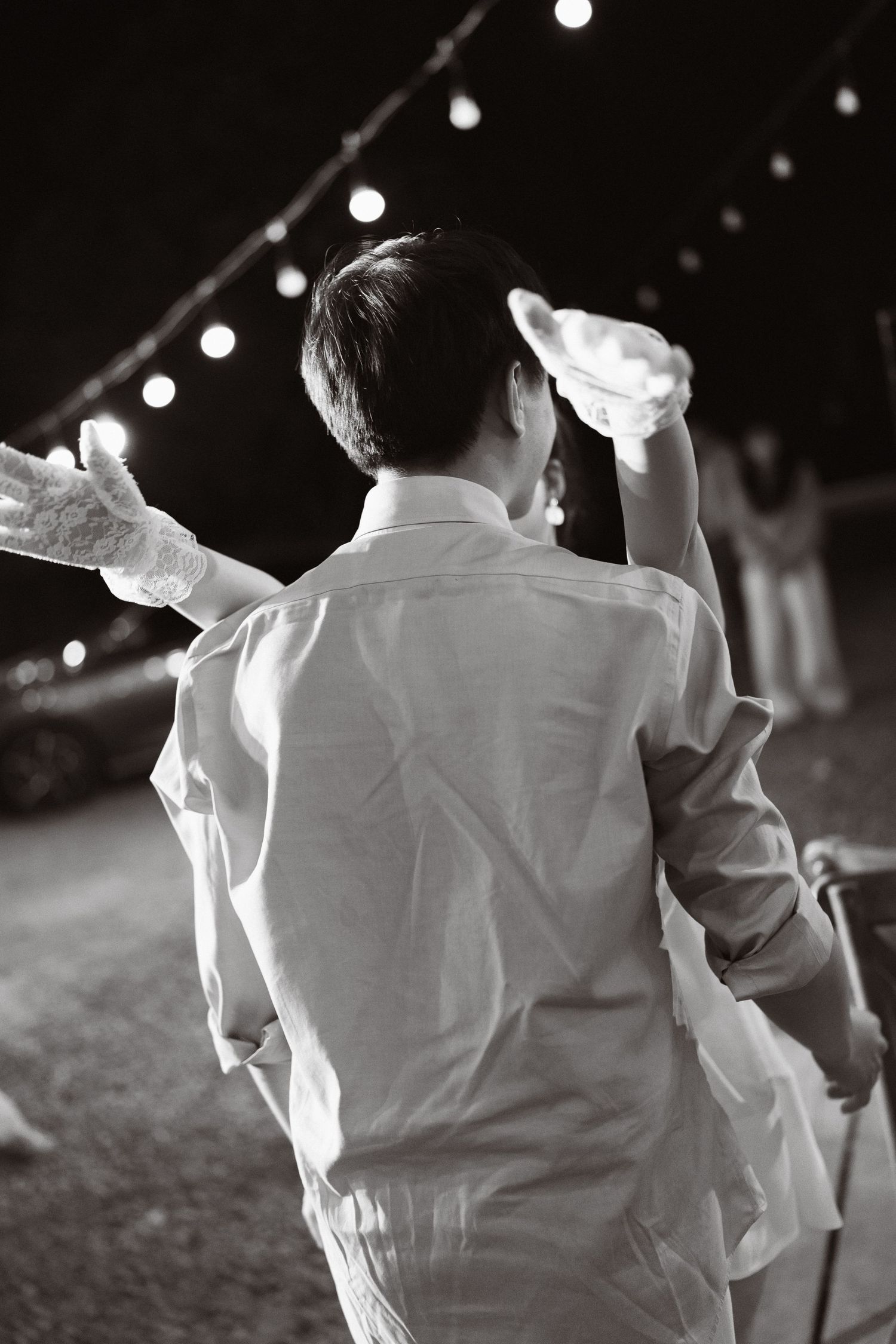 Black and white photo of a man in white shirt holding hat outdoors at night with string lights.
