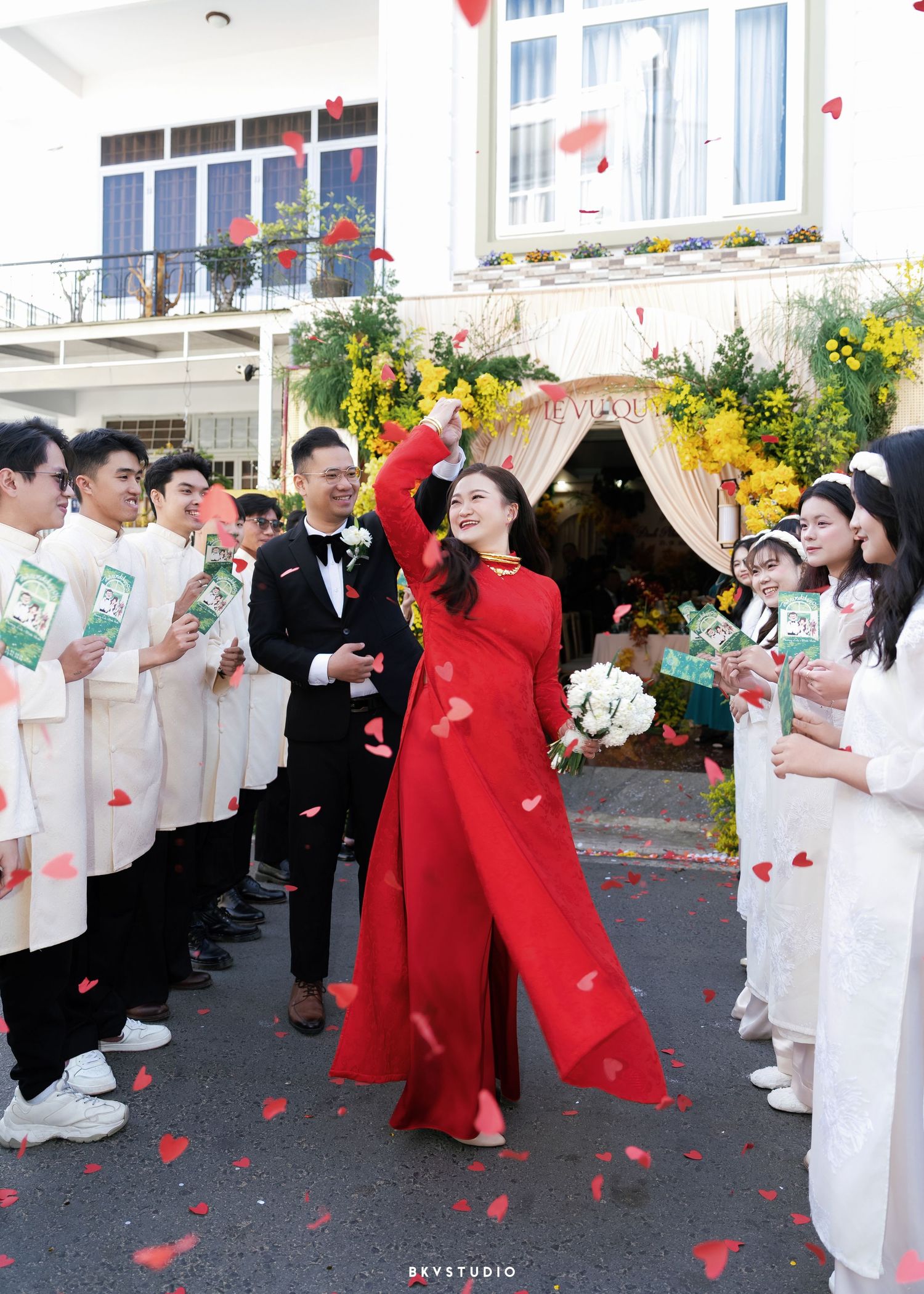 A bride in a red ao dai raises her bouquet joyfully as guests toss rose petals during a traditional Vietnamese wedding ceremony.