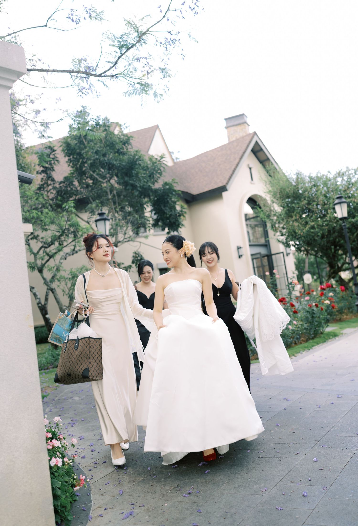 Bride in white strapless gown walks with bridesmaids in black and nude dresses at outdoor wedding venue.