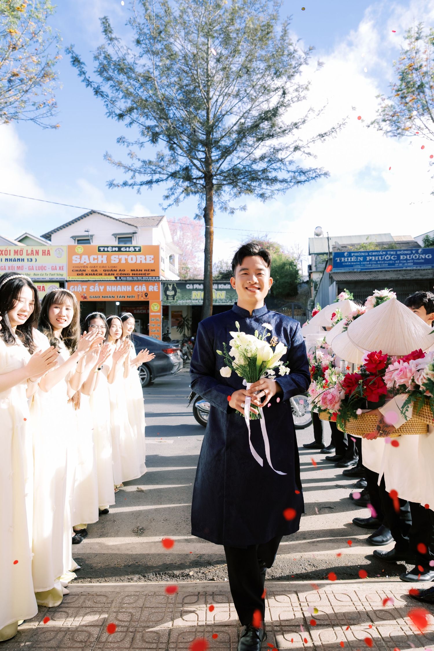 Groom in traditional Vietnamese blue ao dai walks down an outdoor aisle holding a bouquet at his wedding.
