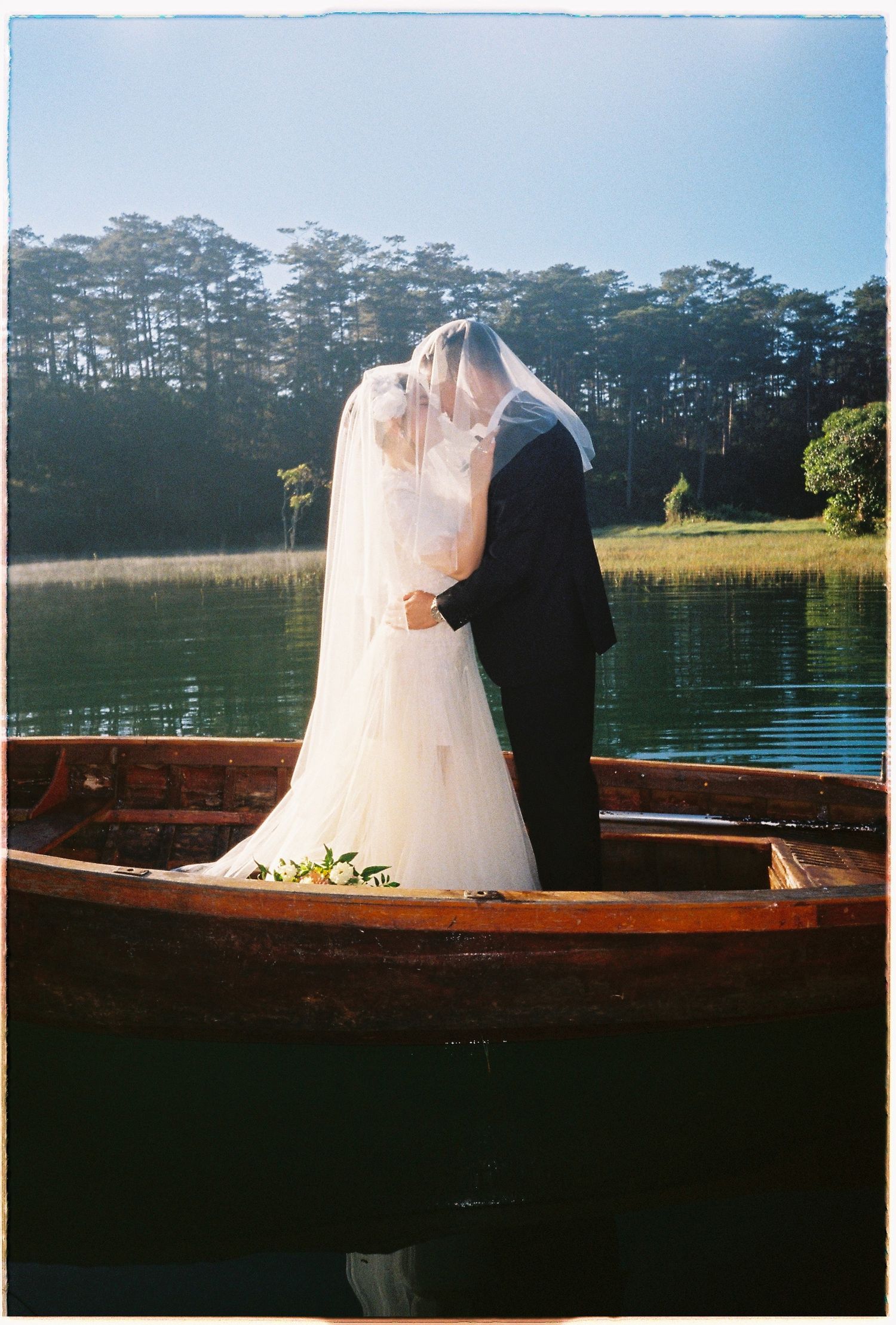 Bride and groom share a romantic kiss under a flowing veil while standing in a wooden boat on a lake.