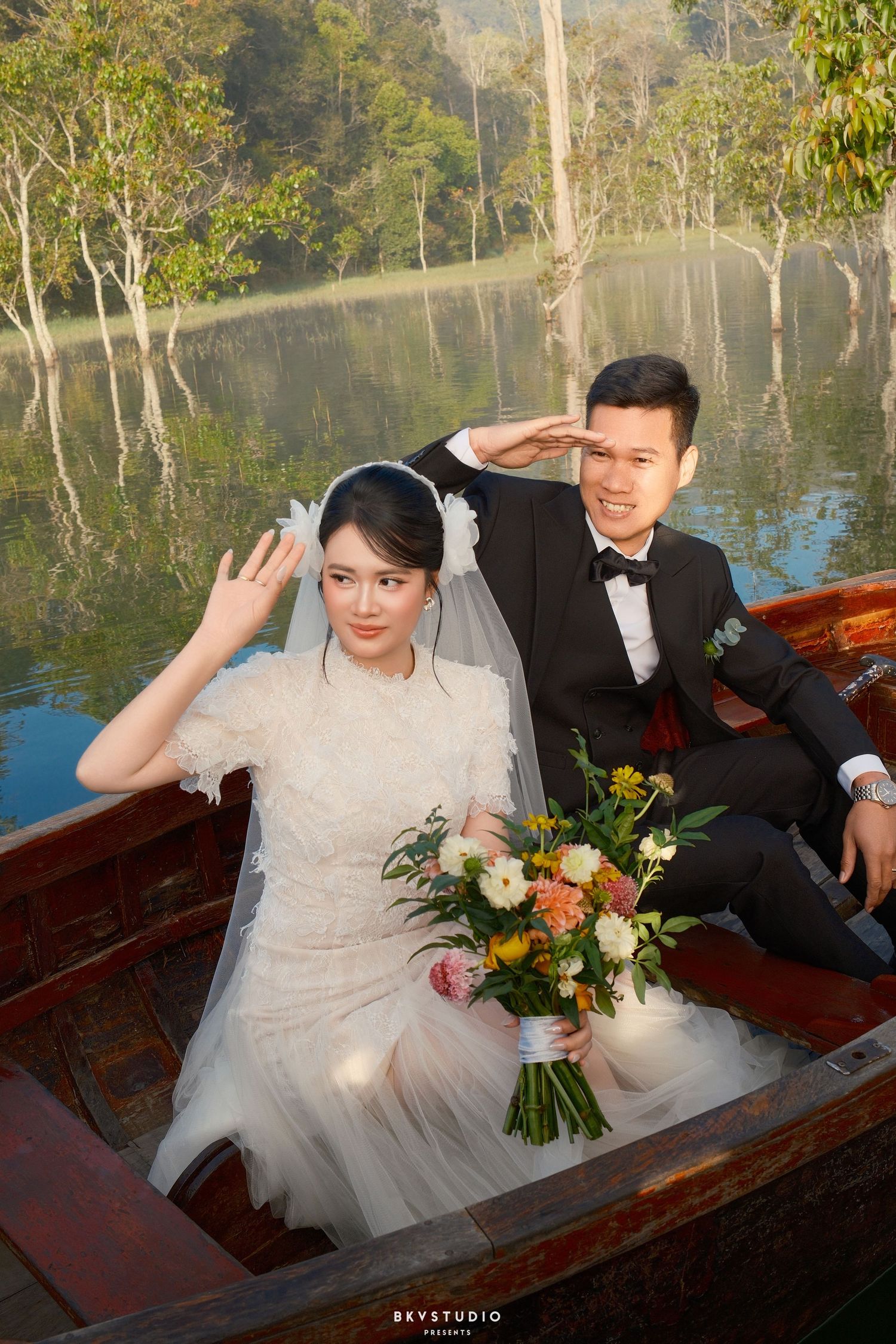 Bride and groom pose playfully in a wooden boat on a calm reflective lake during wedding shoot.