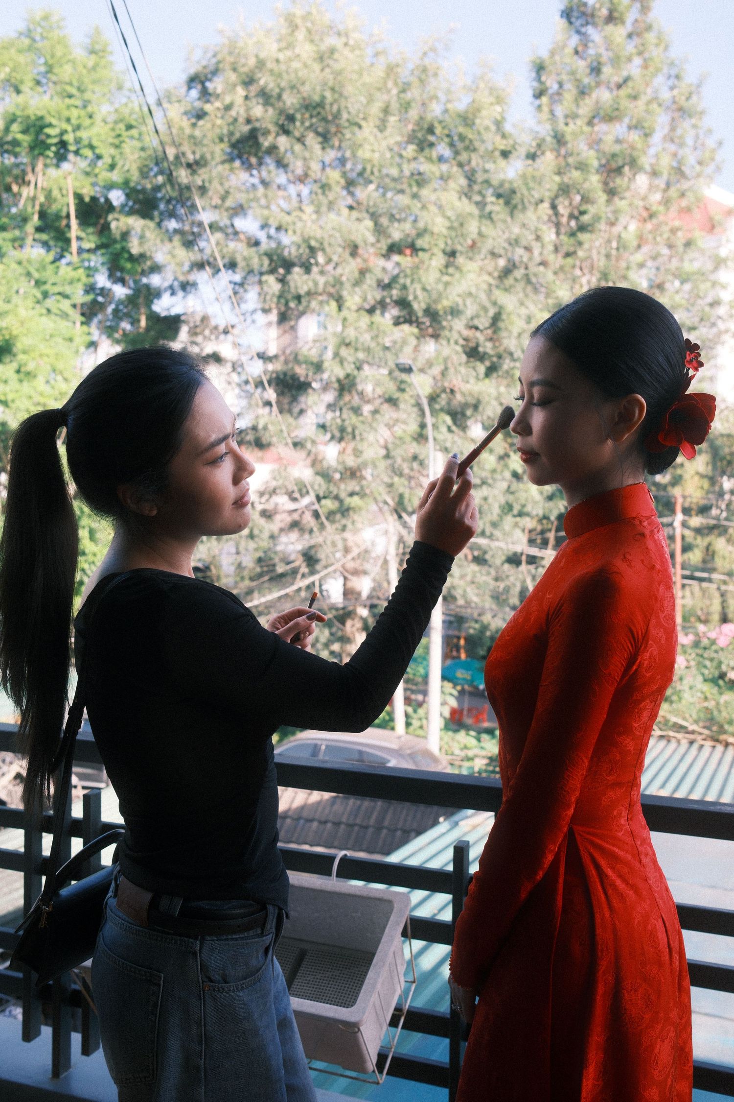 A makeup artist applies foundation to a woman in a red ao dai dress on a balcony with green trees in the background.