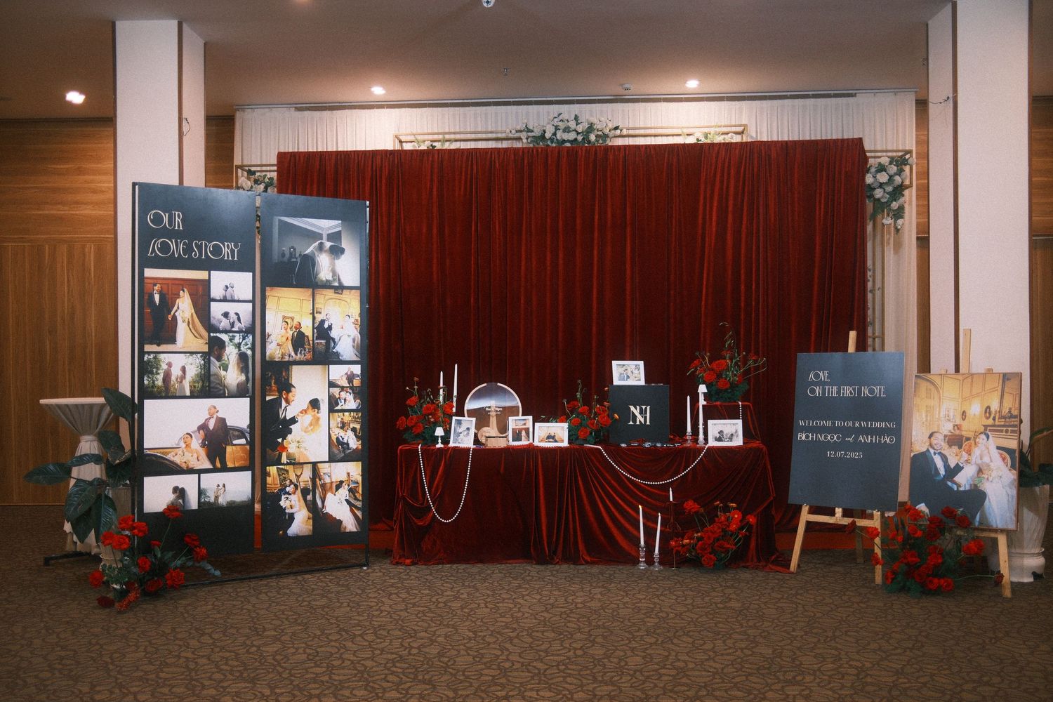Elegant wedding photo display booth with red velvet backdrop, floral arrangements, and framed photos on a decorated table.