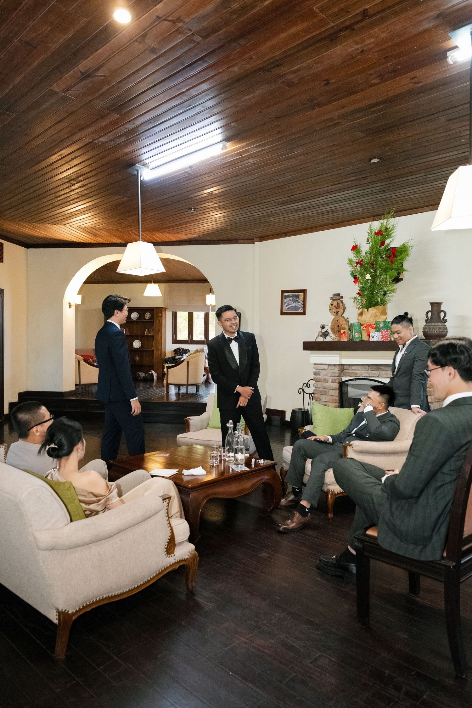 Groom in tuxedo socializing with groomsmen in suits inside a vintage wooden-ceiling venue.