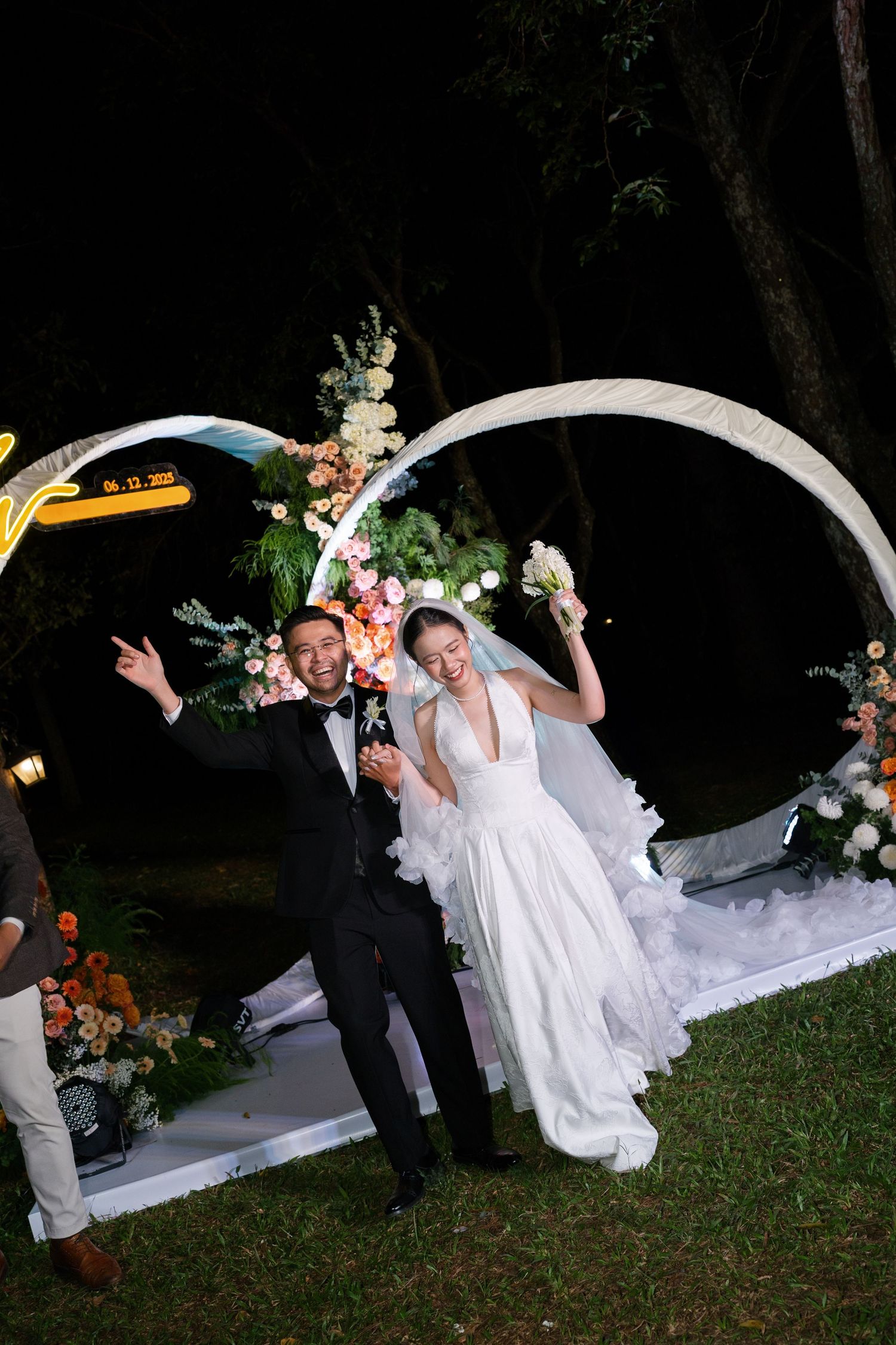 Joyful bride and groom celebrating at night wedding ceremony under floral arch with neon lights.