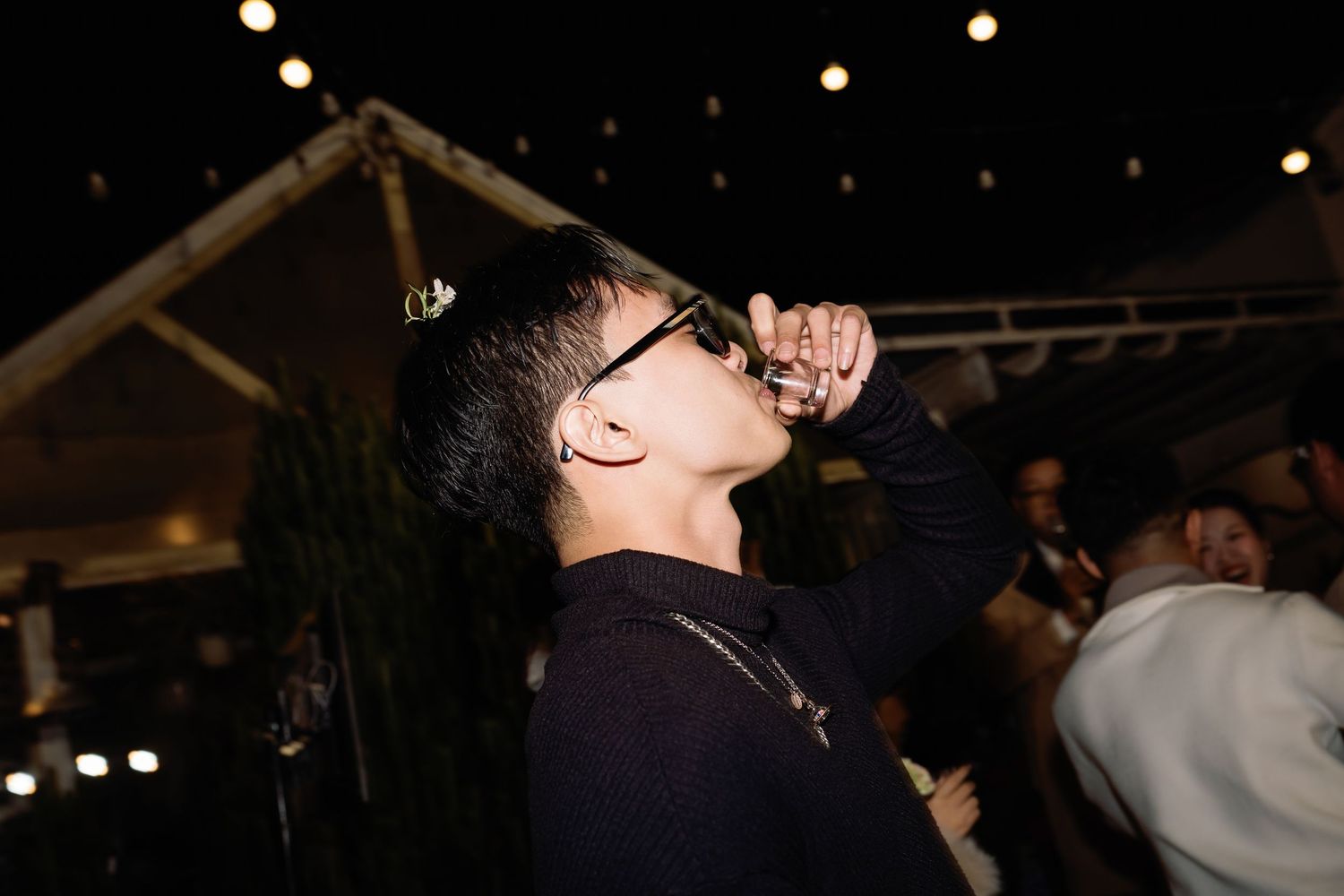 Person in glasses drinking at an outdoor night event with string lights in the background.