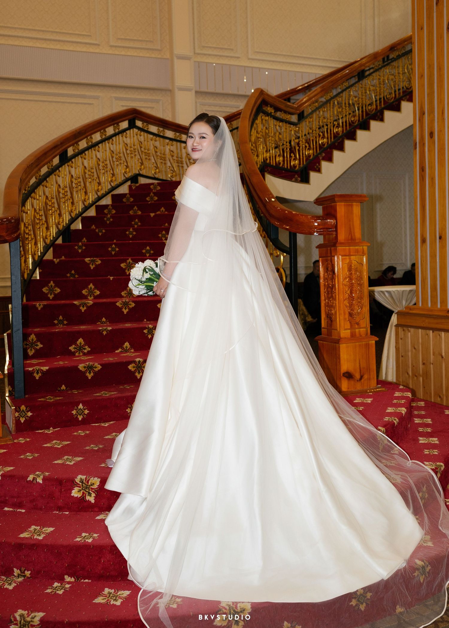 Bride in elegant white ball gown with veil poses on grand staircase with red carpet.