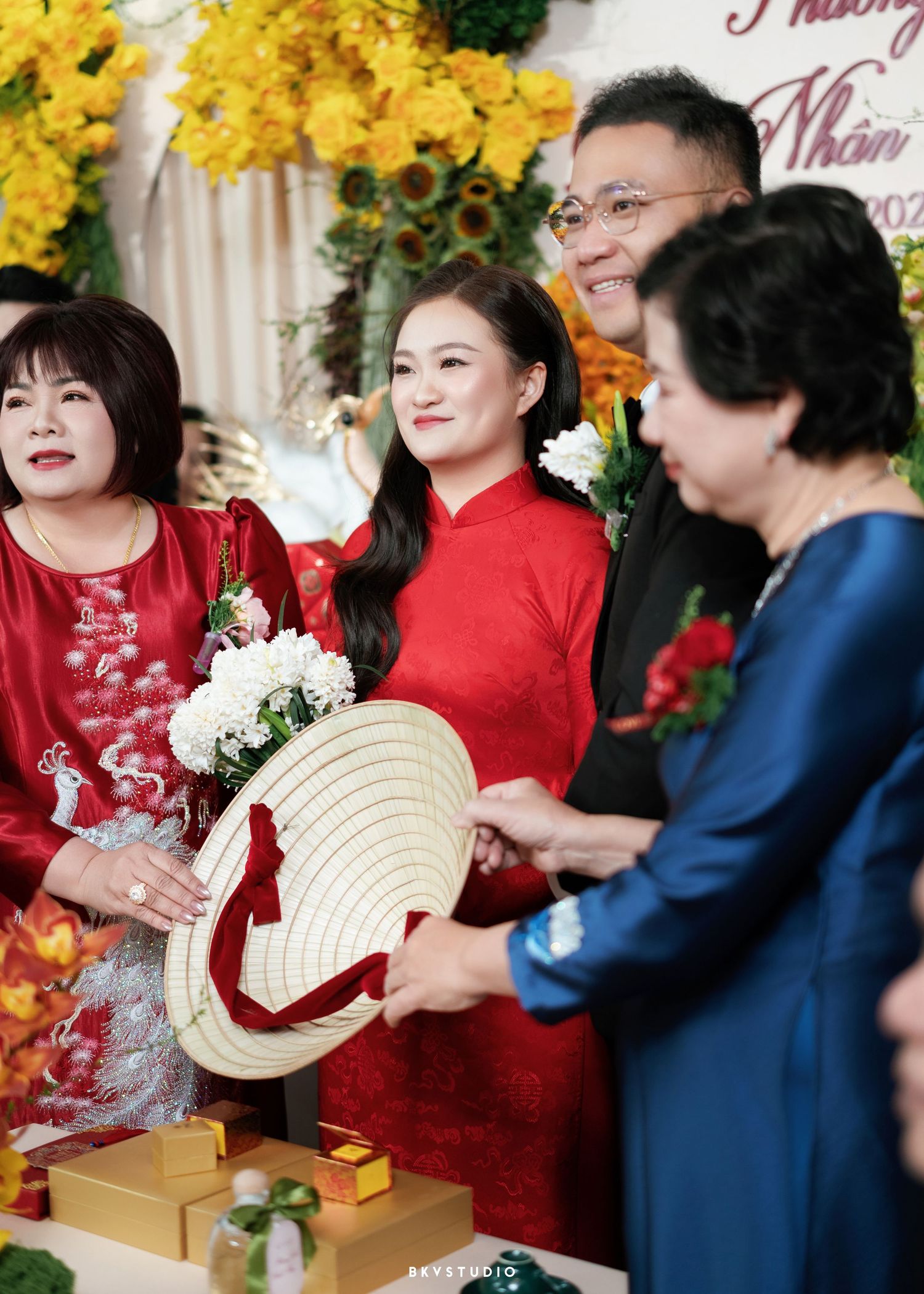 Vietnamese bride in red ao dai receives a traditional conical hat during a wedding engagement ceremony with family members.