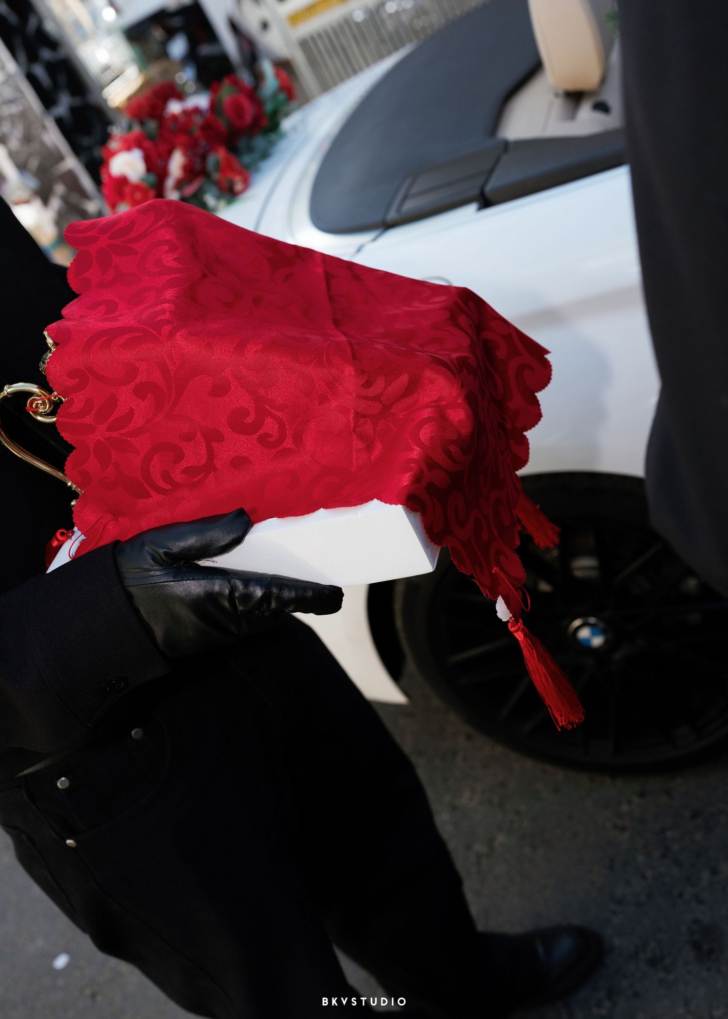 Person in black gloves holding a red embroidered fabric with tassel near a white car.