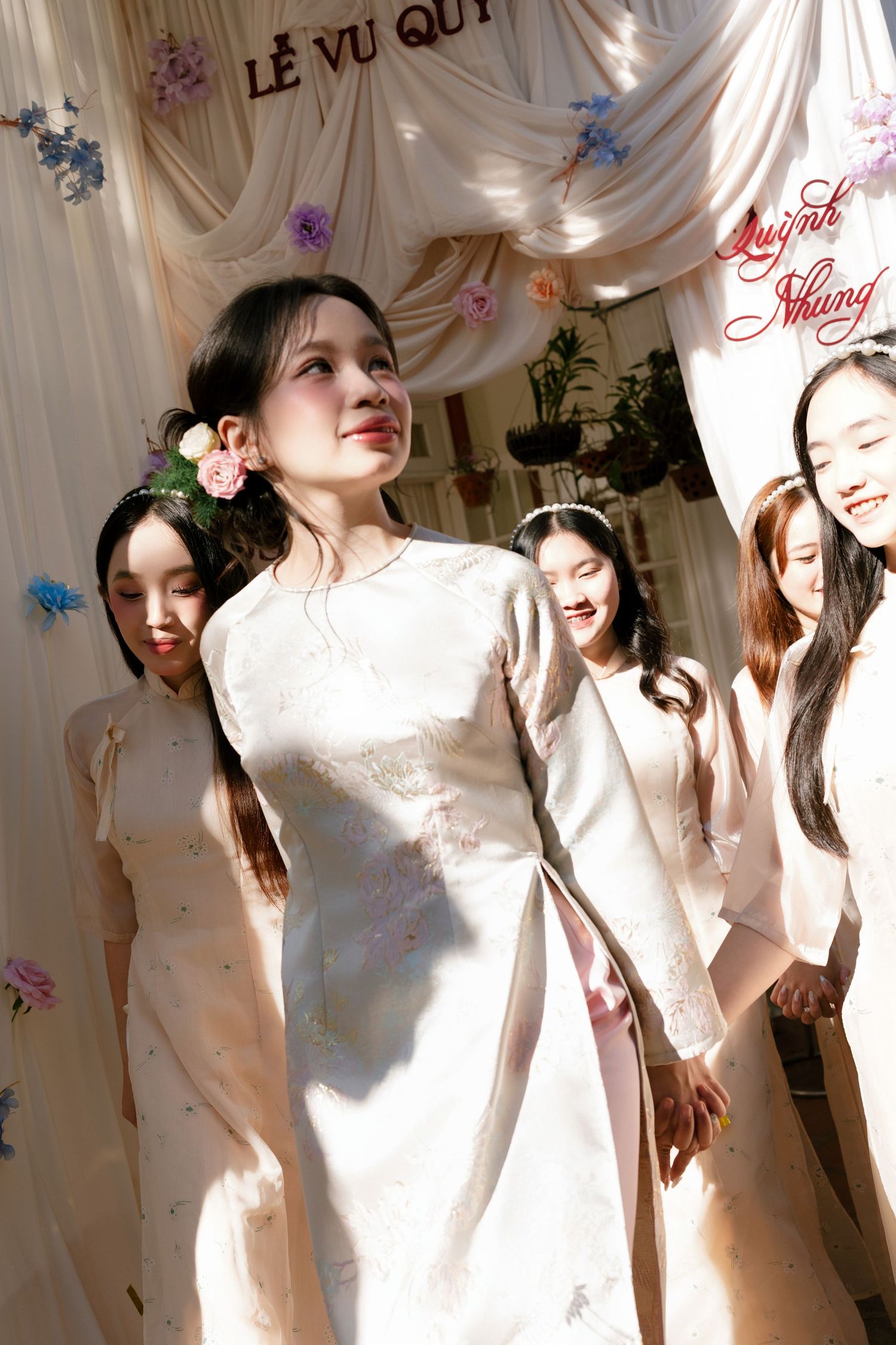 Young women in white ao dai dresses at a Vietnamese wedding ceremony decorated with flowers and draping fabric.