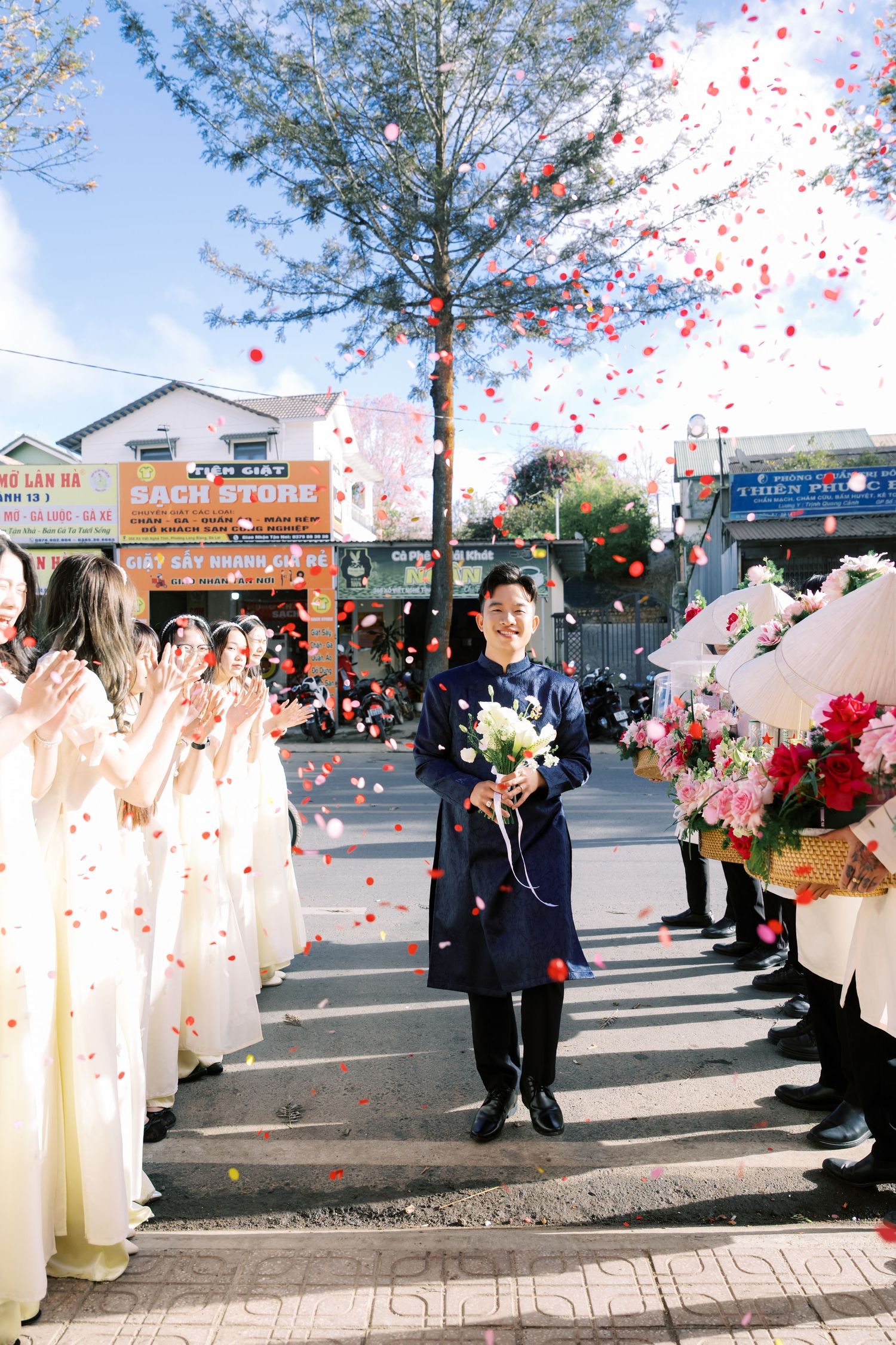 Groom walks through confetti-lined aisle in navy suit holding bouquet at Vietnamese outdoor wedding ceremony.