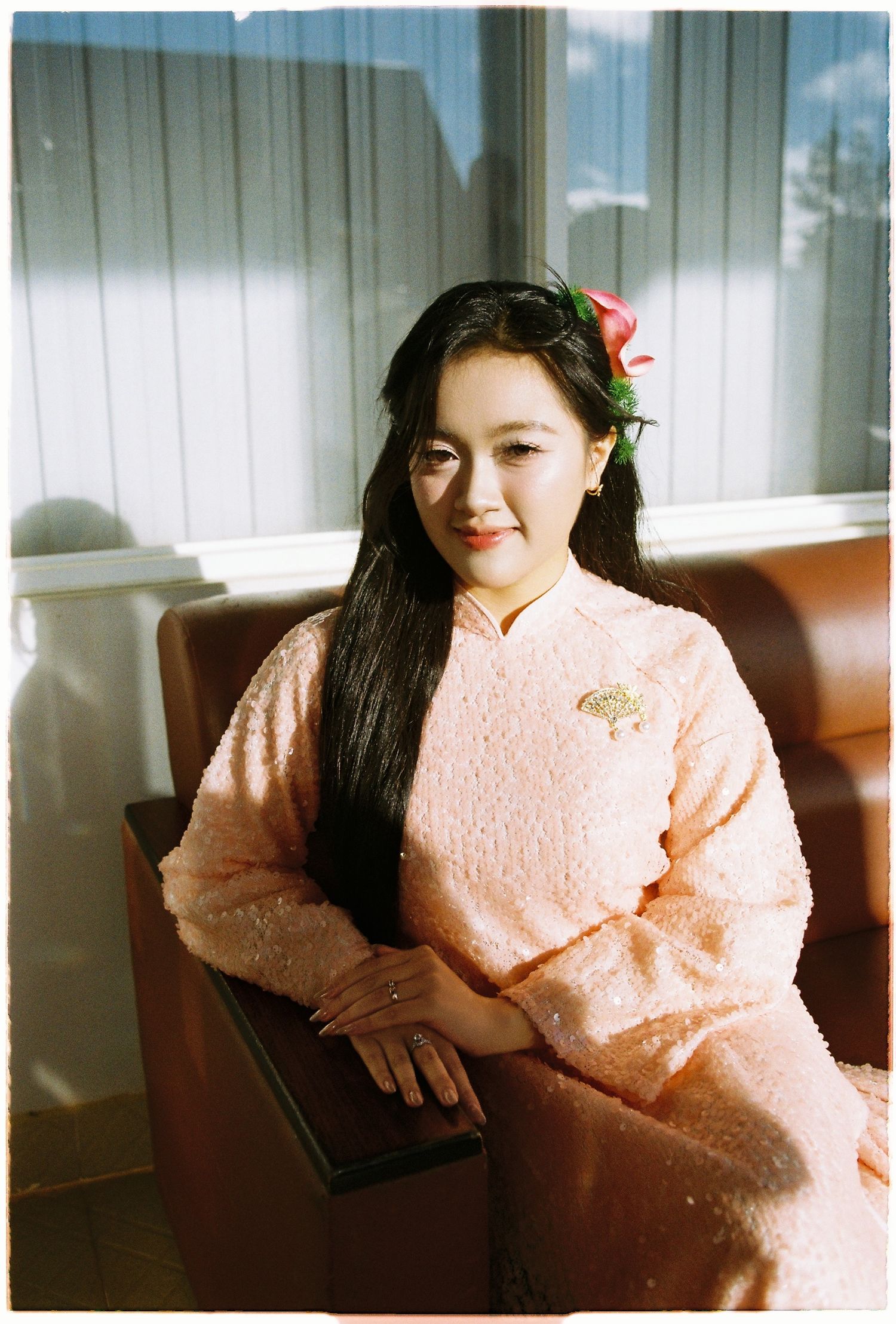 Woman in pink traditional ao dai with floral hair accessory sits in leather chair by window.