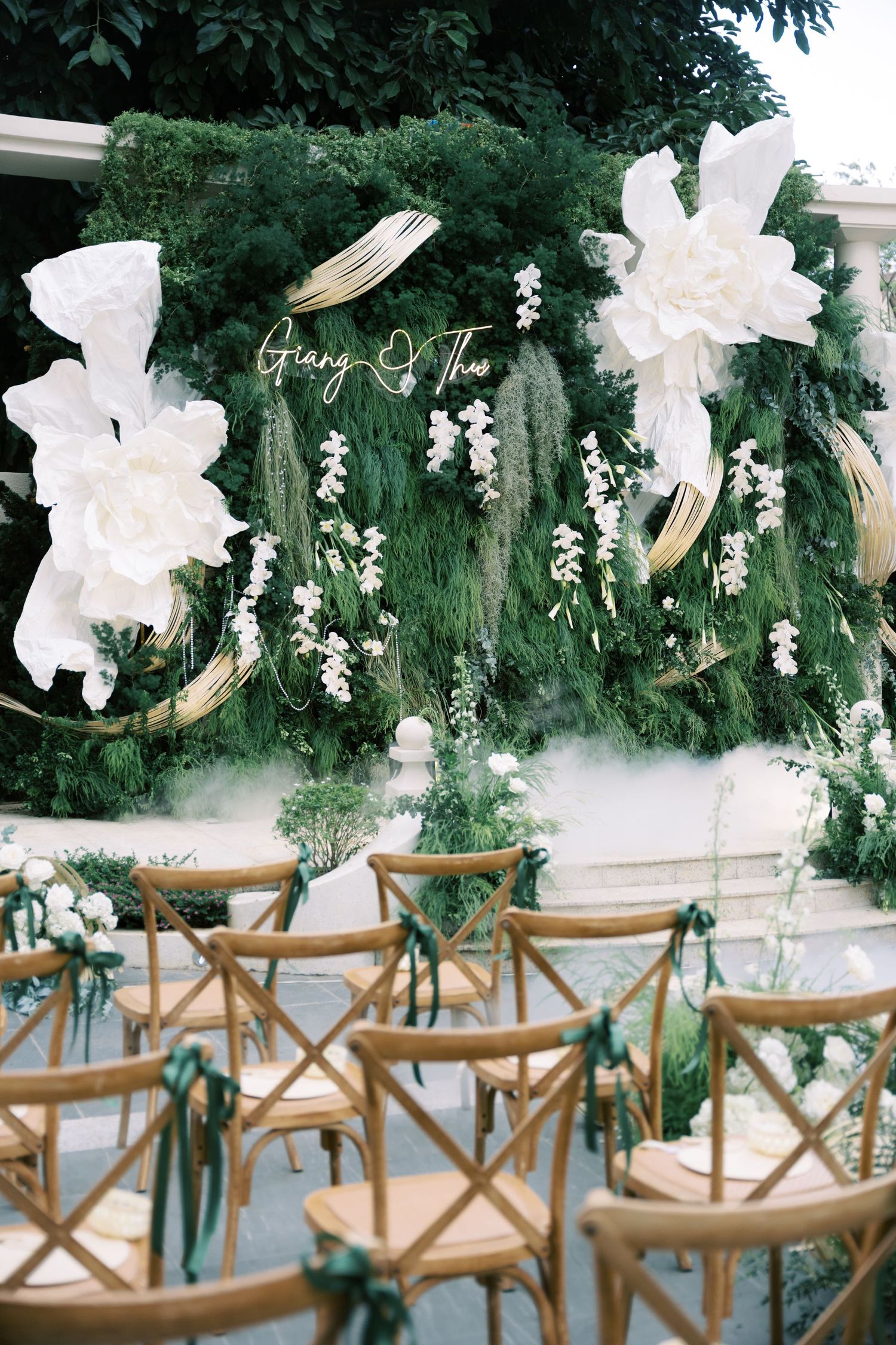 Outdoor wedding ceremony setup with green floral backdrop, wooden cross-back chairs, and white paper flowers.