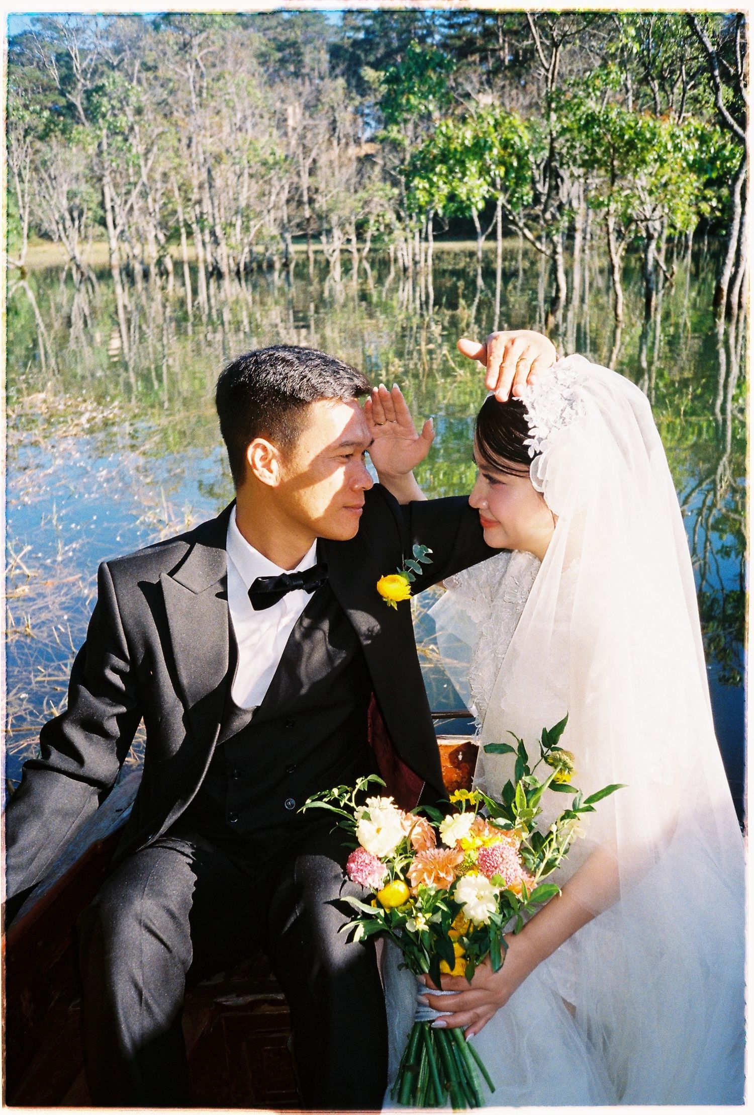 Bride and groom share an intimate forehead-touch moment in a boat by a reflective forest lake.