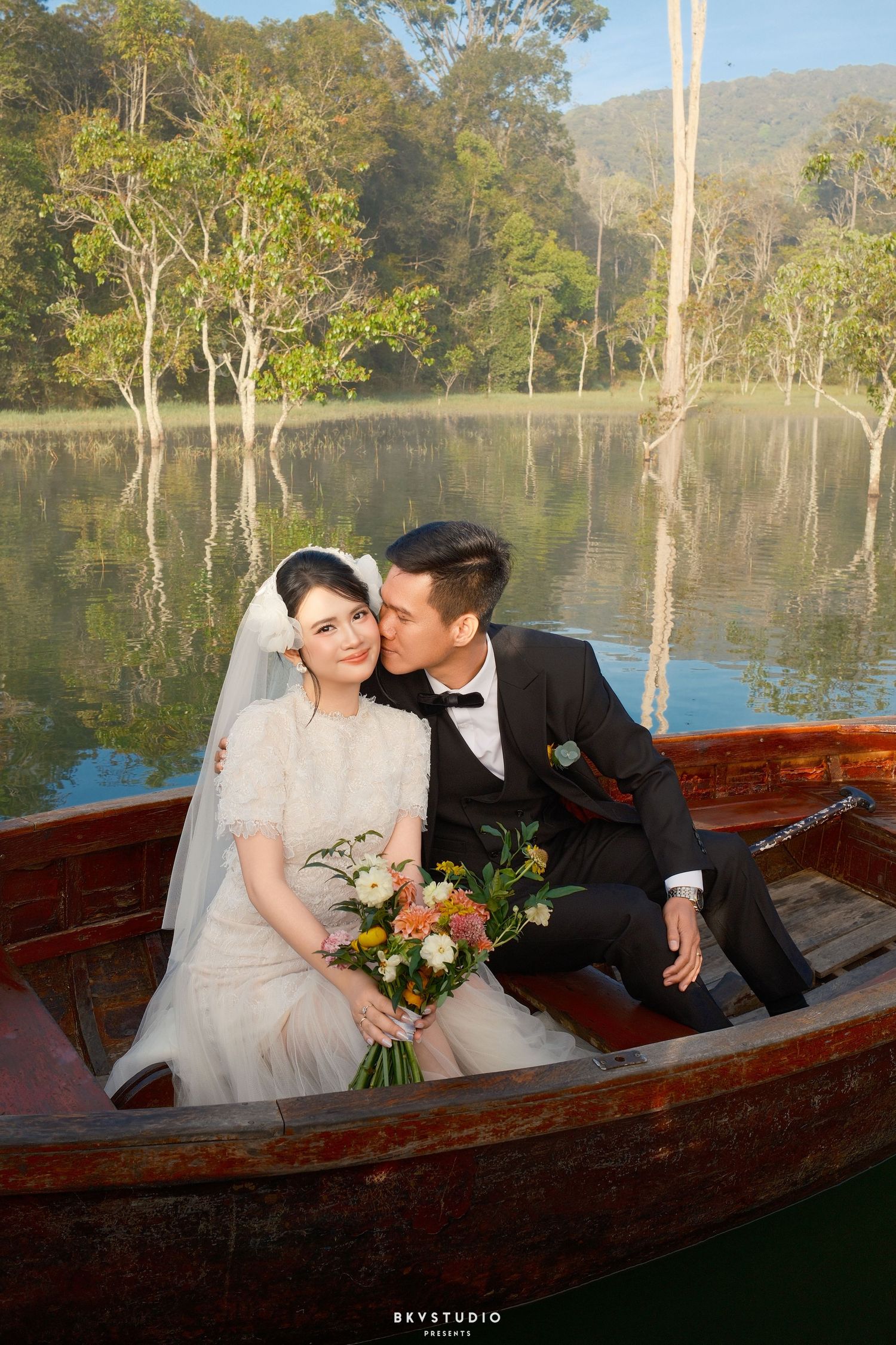 Bride and groom share a romantic kiss on a wooden boat on a calm lake surrounded by trees.