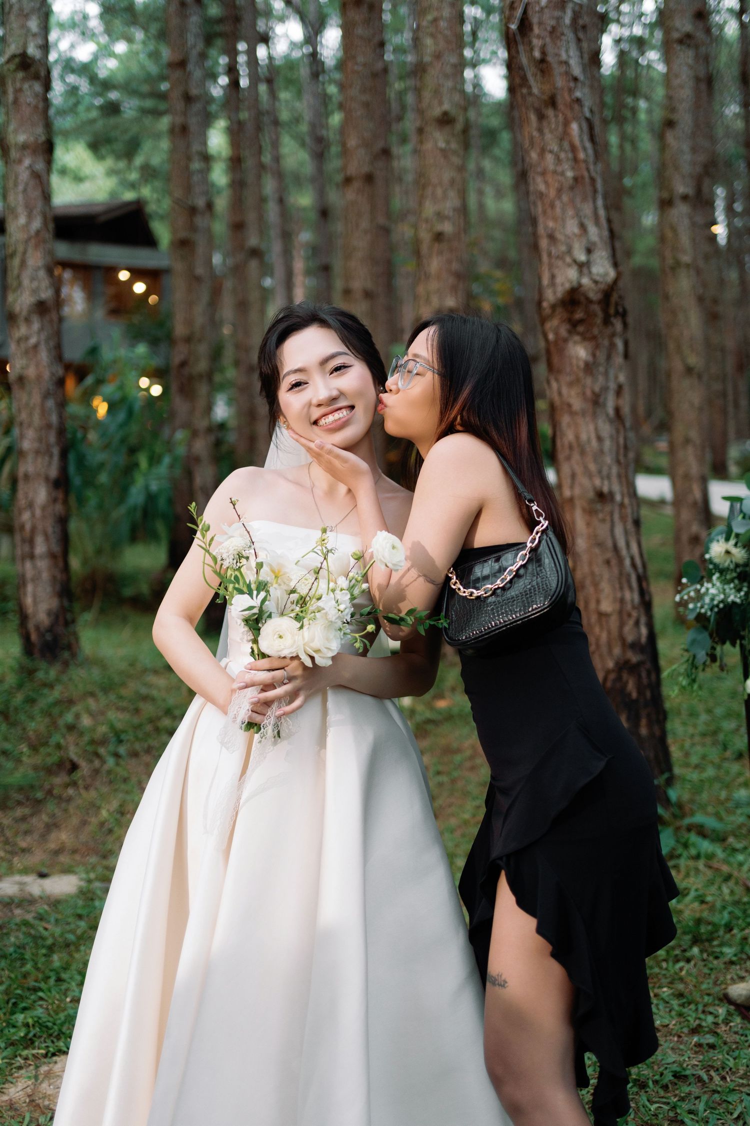 Smiling Asian bride in white strapless gown receives a kiss on the cheek from a friend in a pine forest.