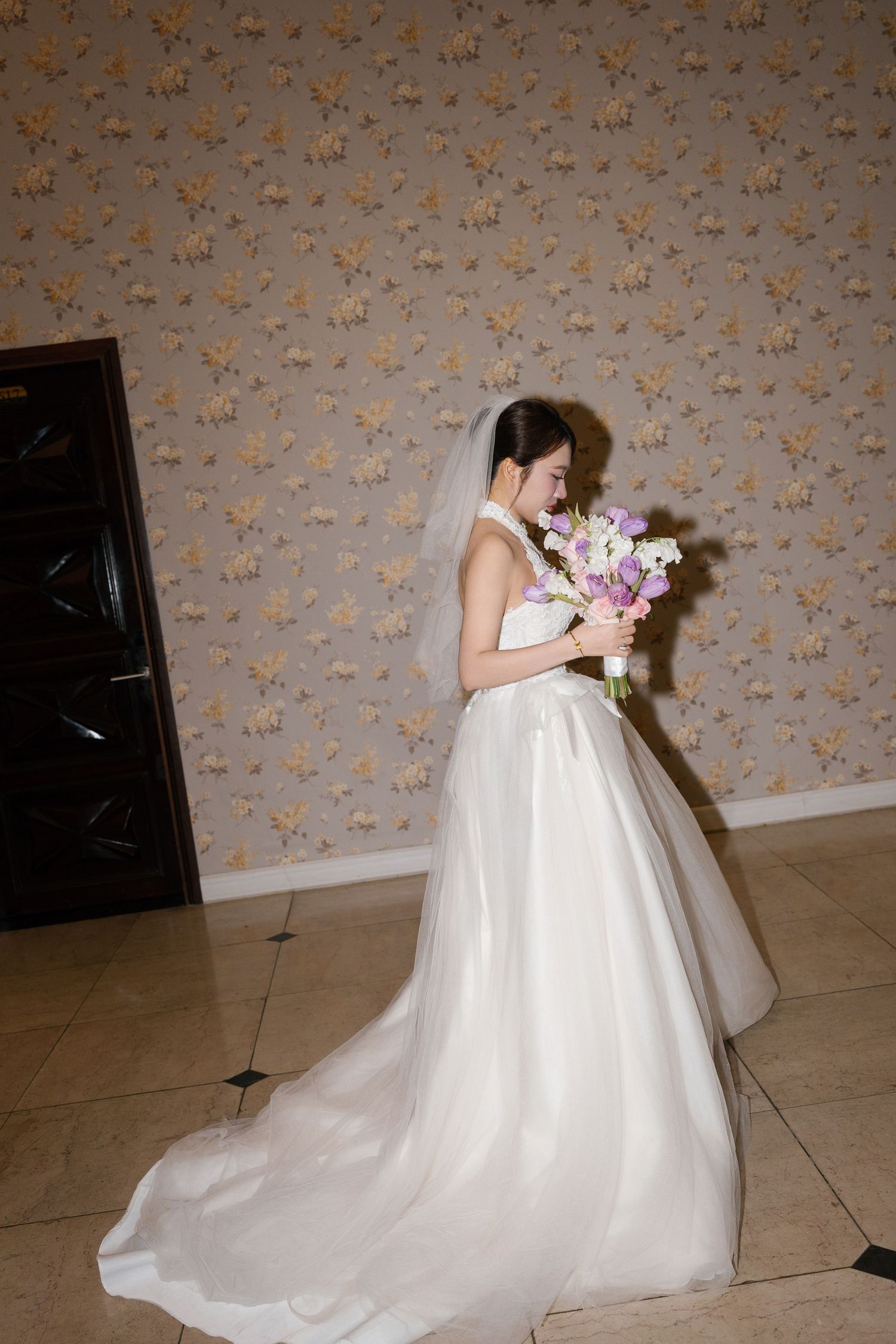 Elegant bride in white ball gown with veil holds purple and white bouquet, posing indoors against floral wallpaper.