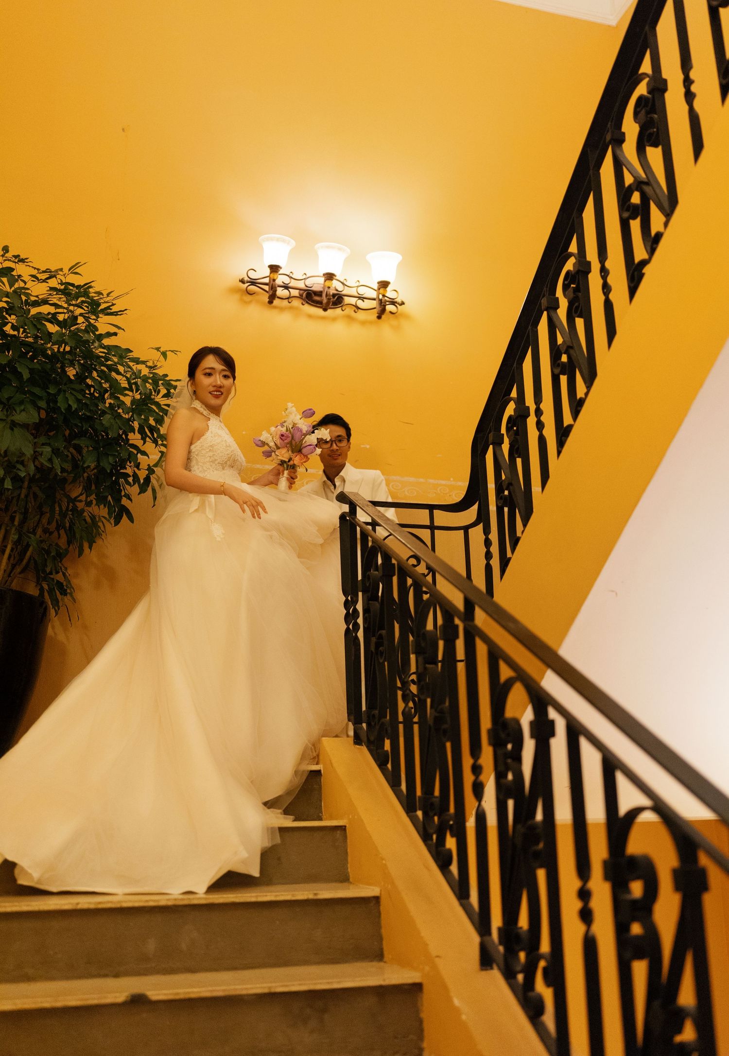 Bride and groom pose on elegant staircase with yellow walls and black iron railing during wedding celebration.