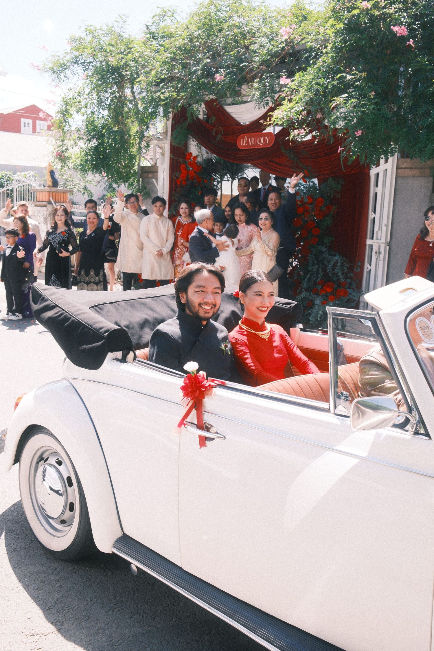 Smiling couple in traditional red attire rides a decorated white vintage VW Beetle convertible at a festive wedding celebration.