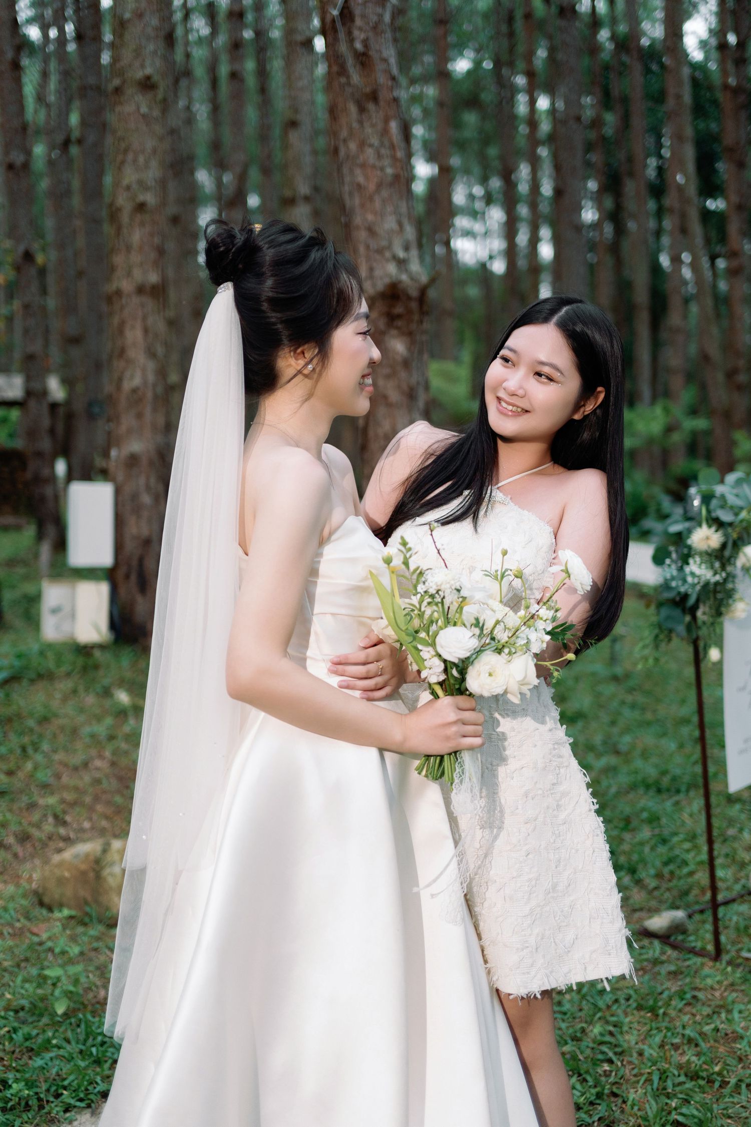 Two Asian women in white dresses embrace and smile at each other during an outdoor forest wedding ceremony.