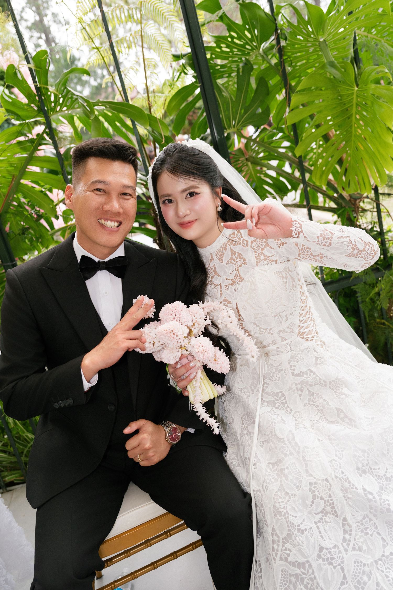 Smiling bride and groom in lace wedding dress posing together in a tropical greenhouse setting.