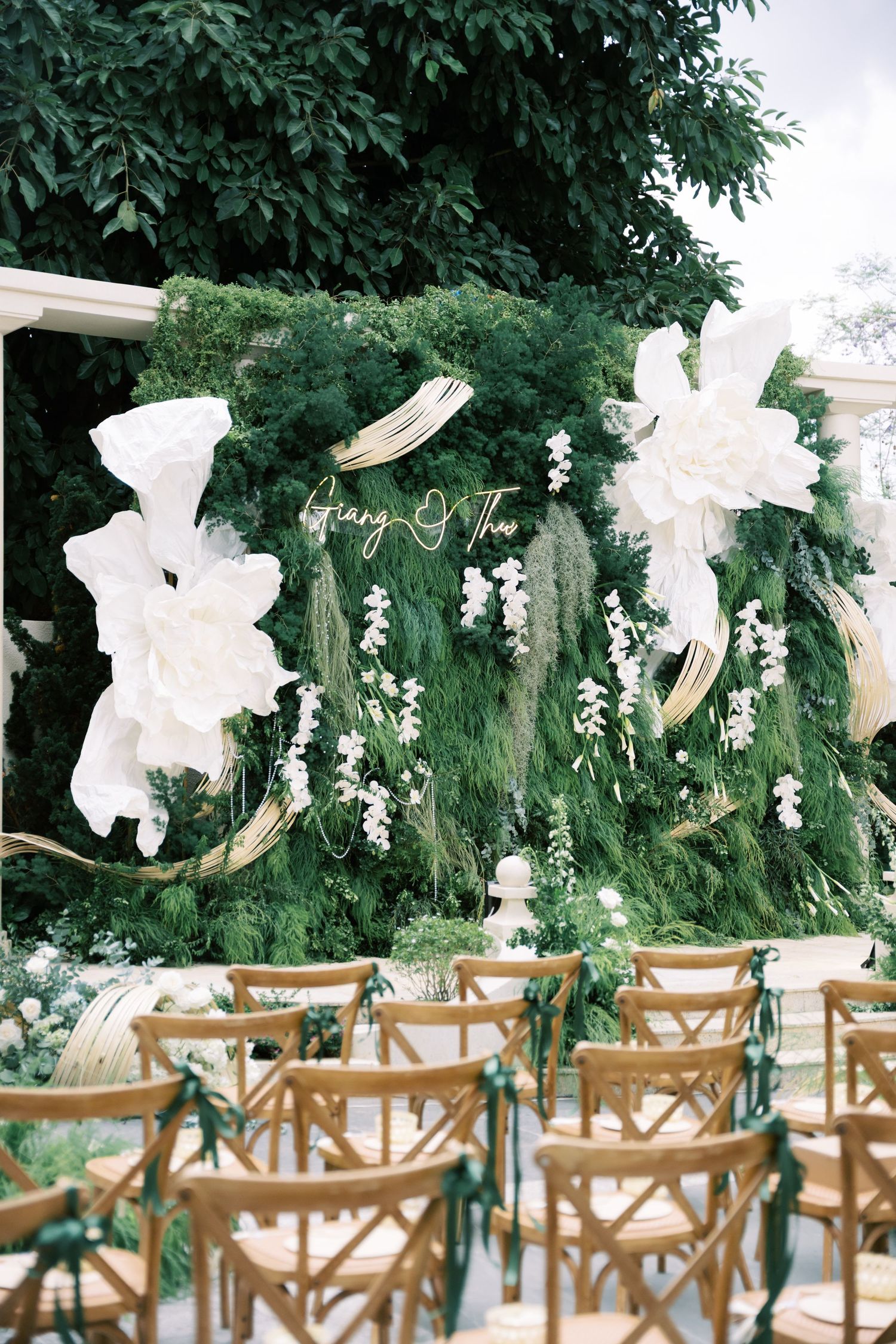 Lush green floral wedding ceremony backdrop with white paper flowers, orchids, and wooden cross-back chairs.