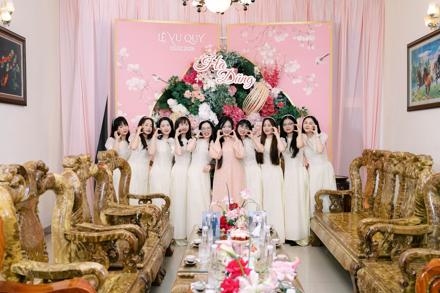 A group of women in white ao dai dresses pose together at a Vietnamese wedding celebration with floral decorations.