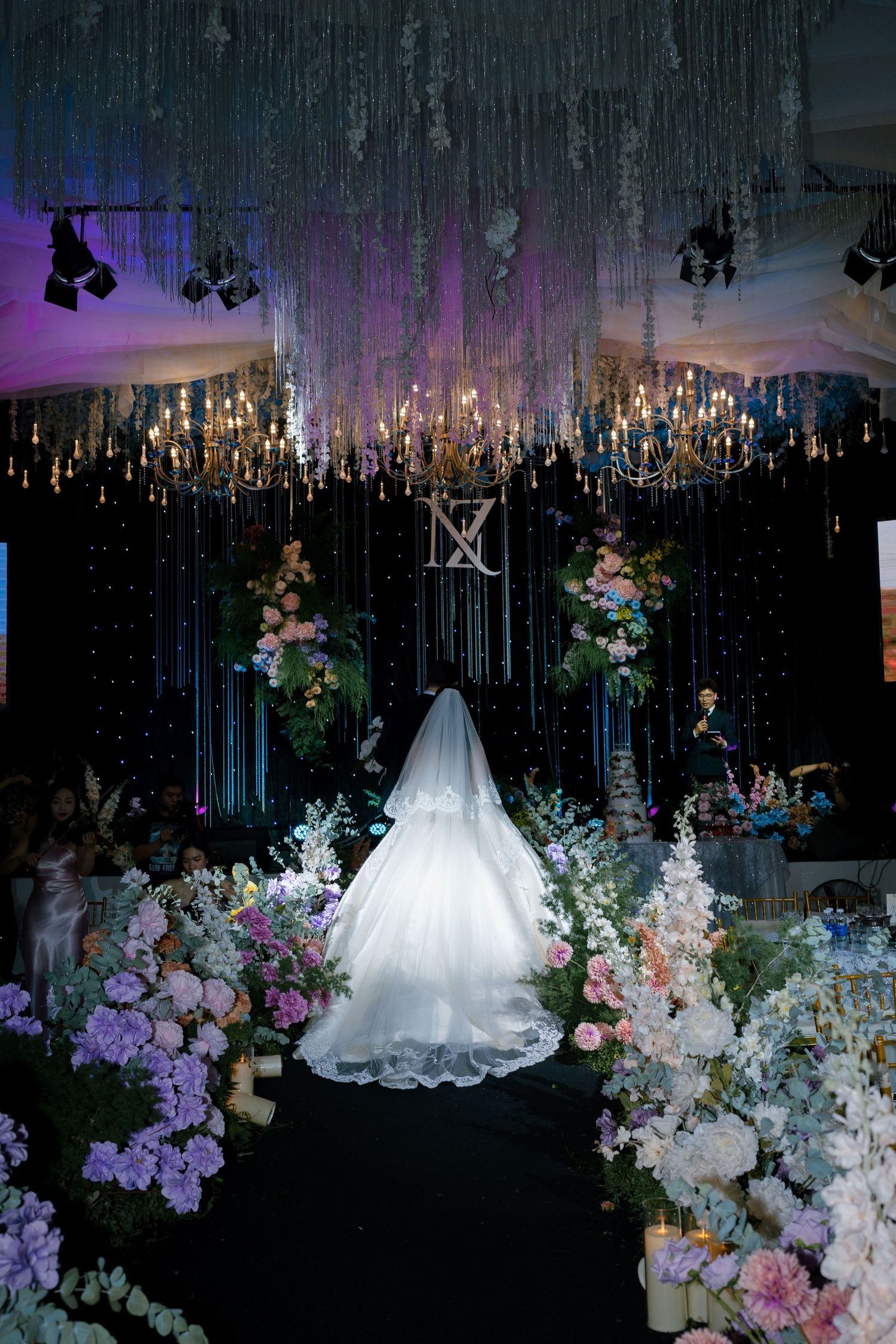Bride in white gown and veil walks down floral aisle with chandeliers and purple lighting at elegant wedding venue.