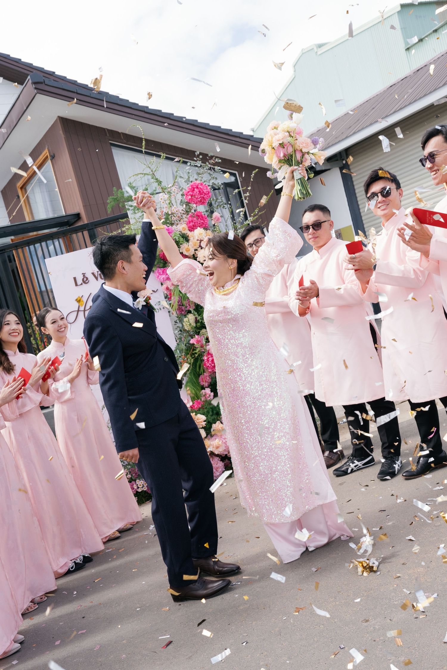 Vietnamese bride and groom celebrating with confetti as wedding party cheers outdoors.