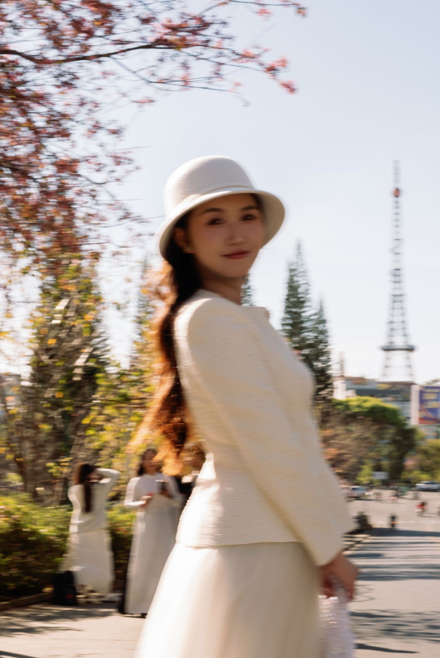 Young woman in white outfit and bucket hat poses outdoors near trees with a tower in the background.