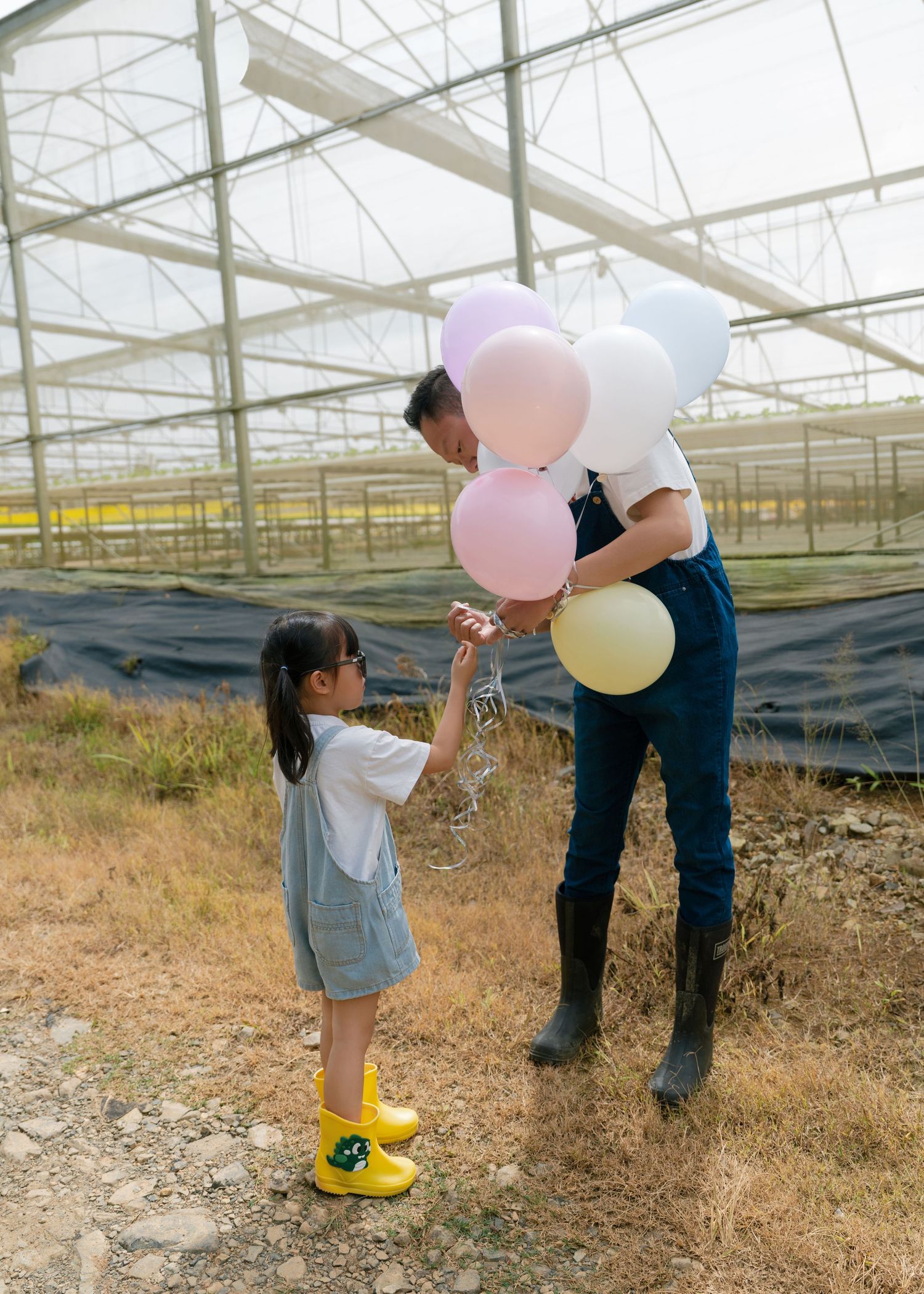 Adult giving colorful pastel balloons to a young girl in yellow boots near a greenhouse on a farm.