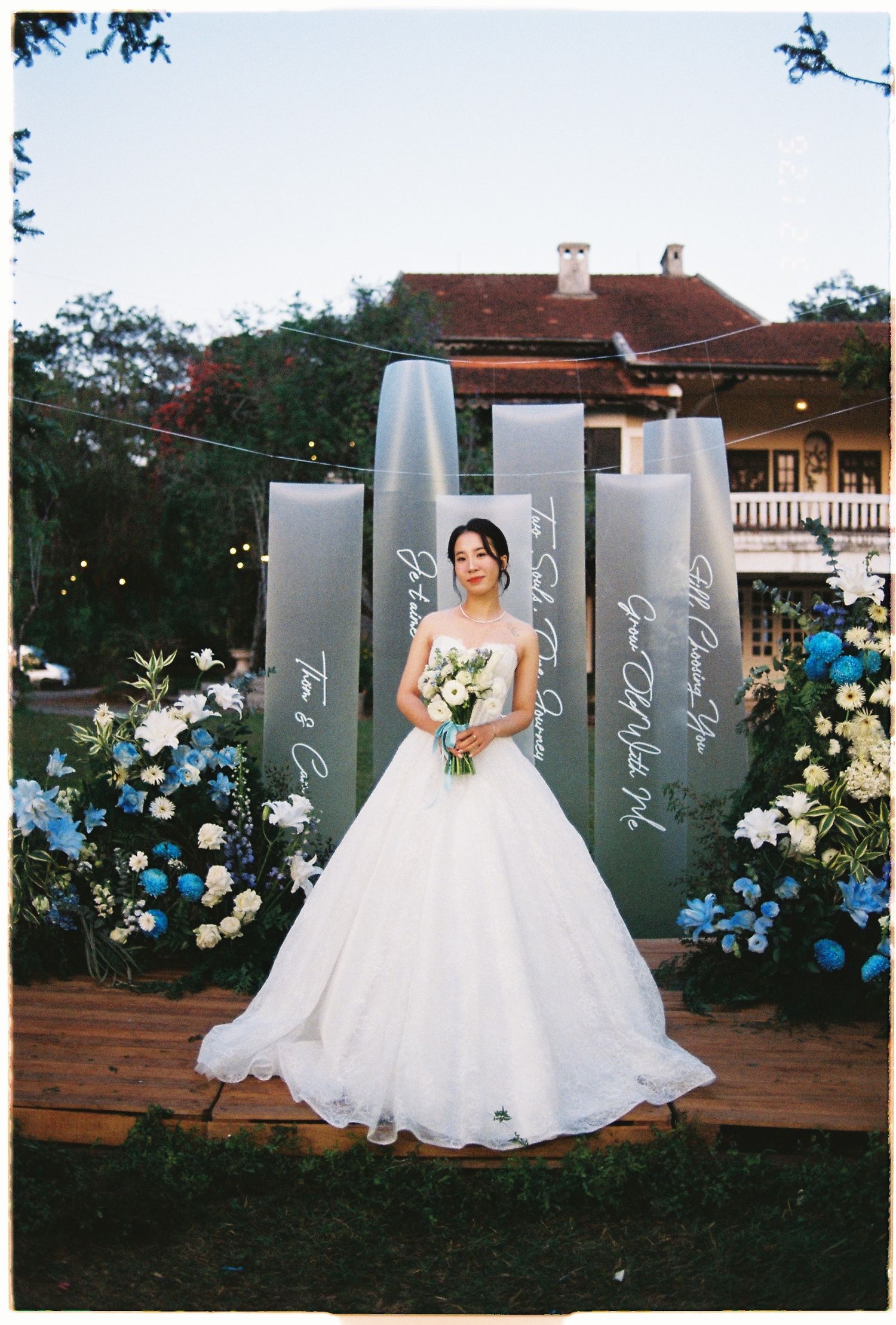 Bride in a white ball gown holding a bouquet stands before a floral backdrop at an outdoor wedding venue.