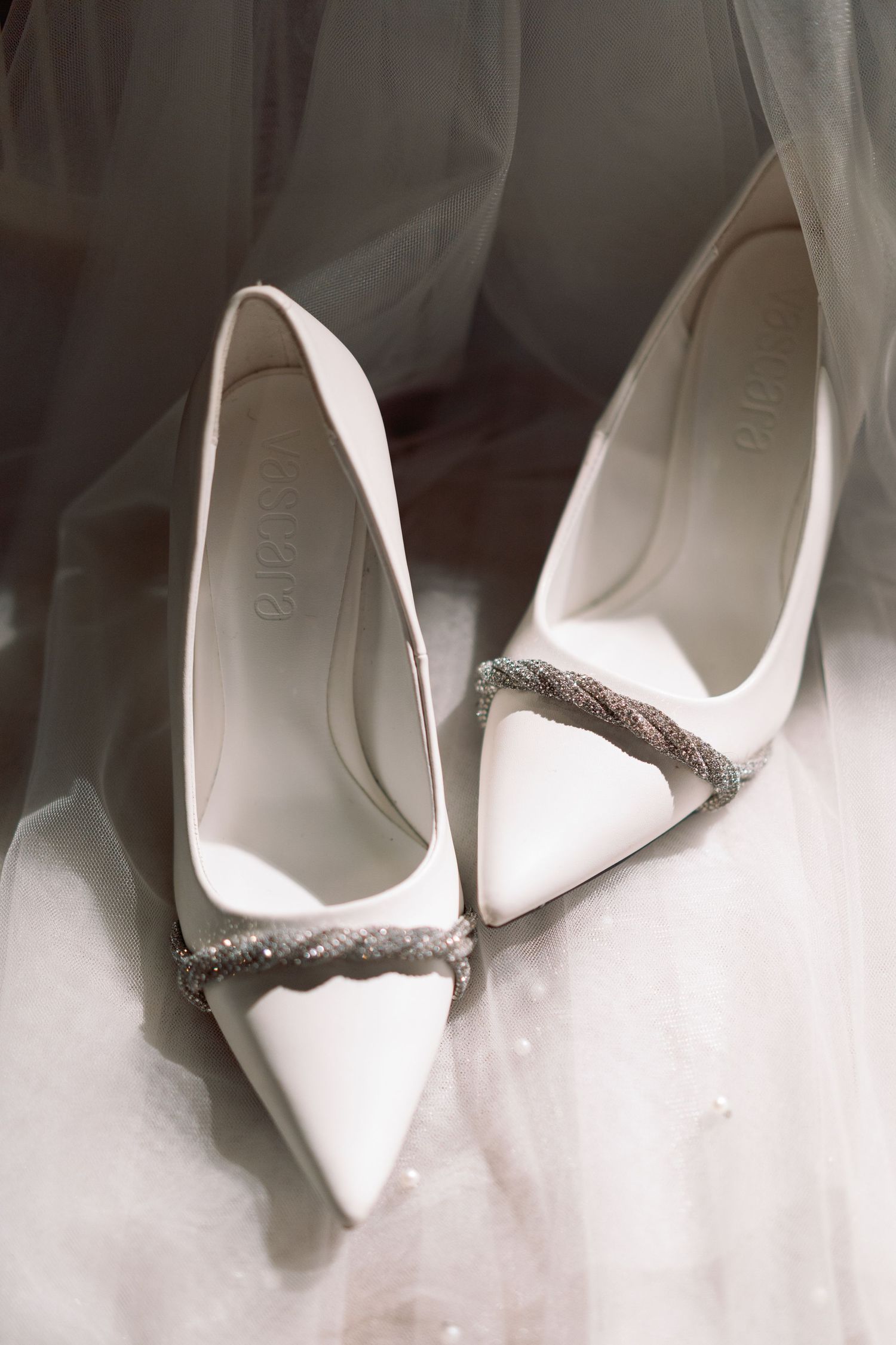 Elegant white pointed-toe bridal heels with crystal embellishment resting on wedding veil.