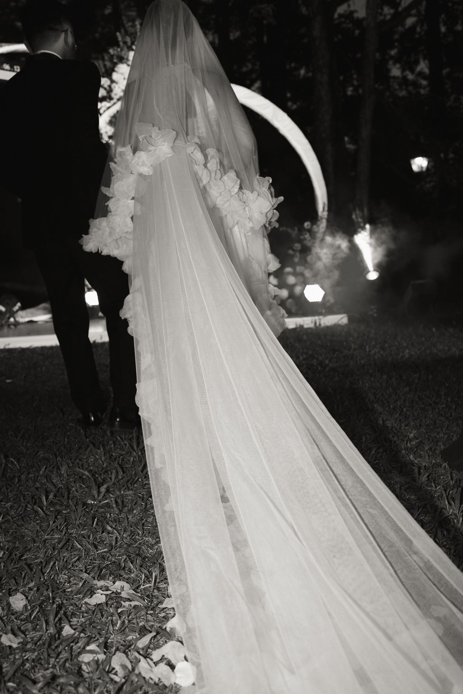Black and white photo of bride with long floral-trimmed cathedral veil trailing on grass at night wedding.