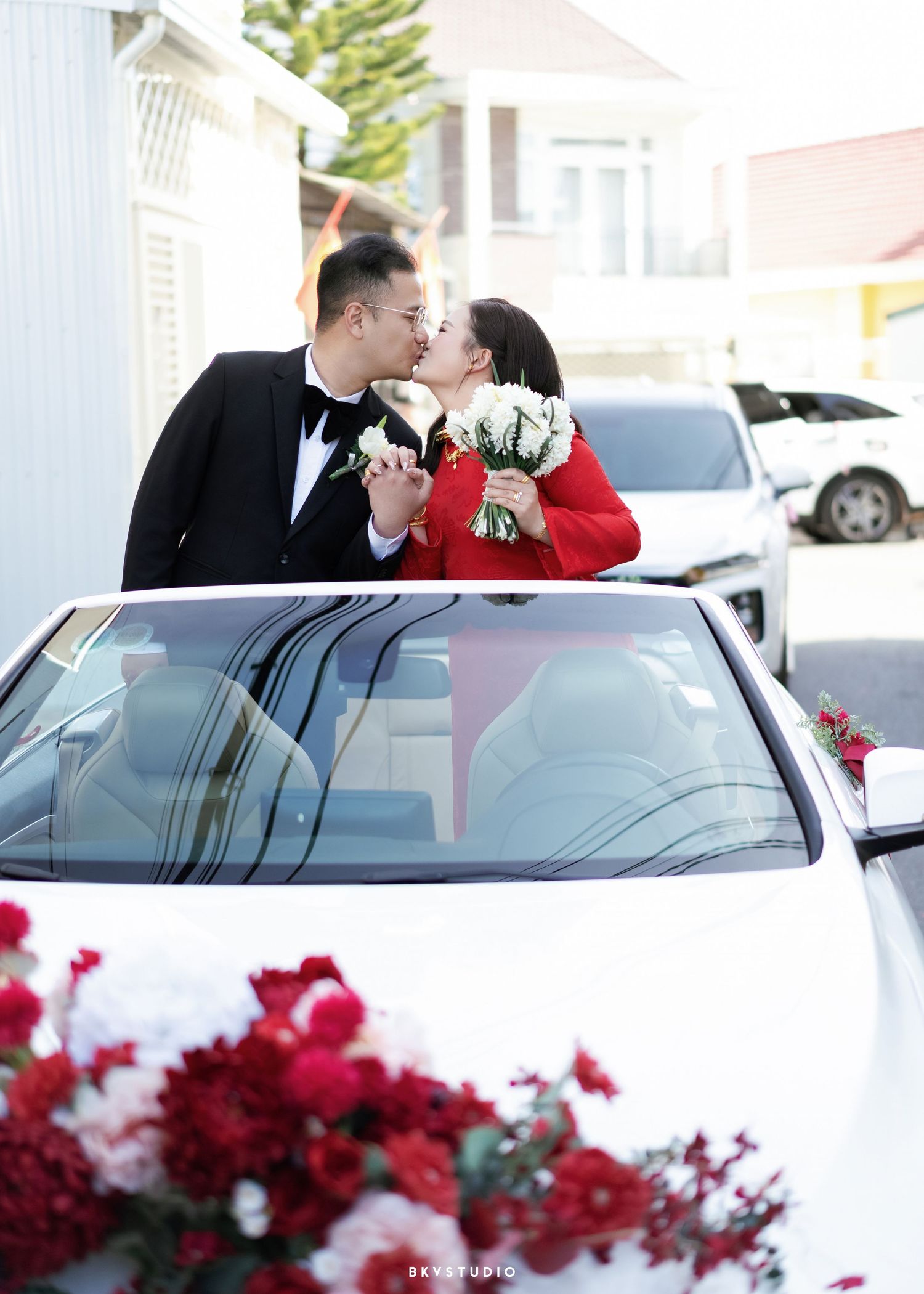 A couple shares a romantic kiss beside a white wedding car adorned with red and white flowers.