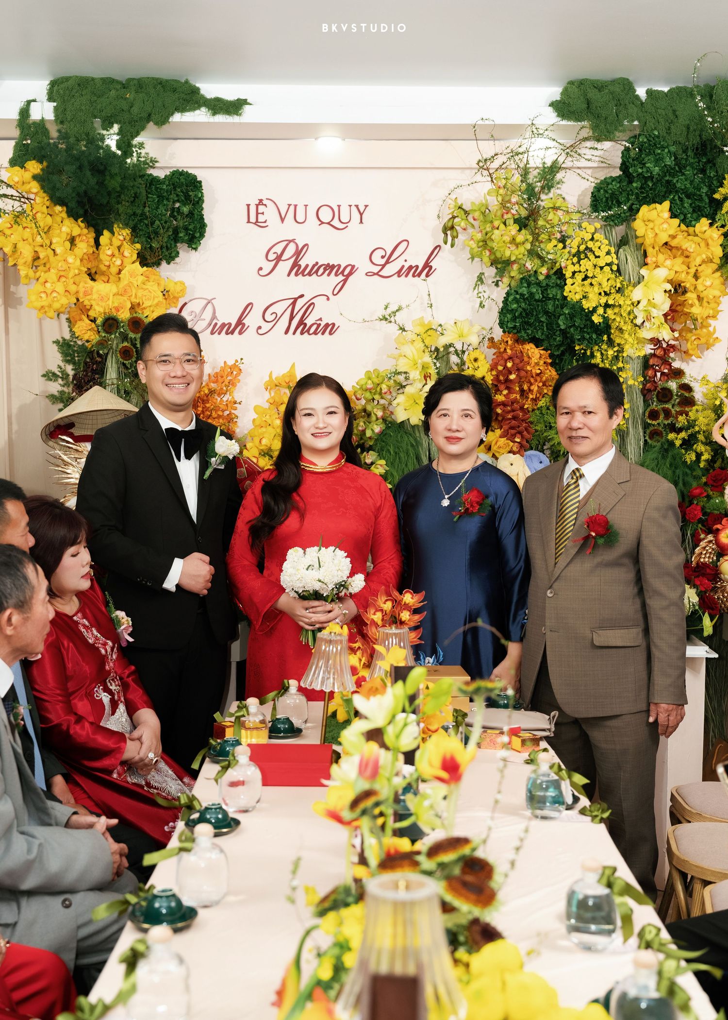 Vietnamese traditional wedding ceremony with couple in ao dai attire, surrounded by family amid floral decorations.