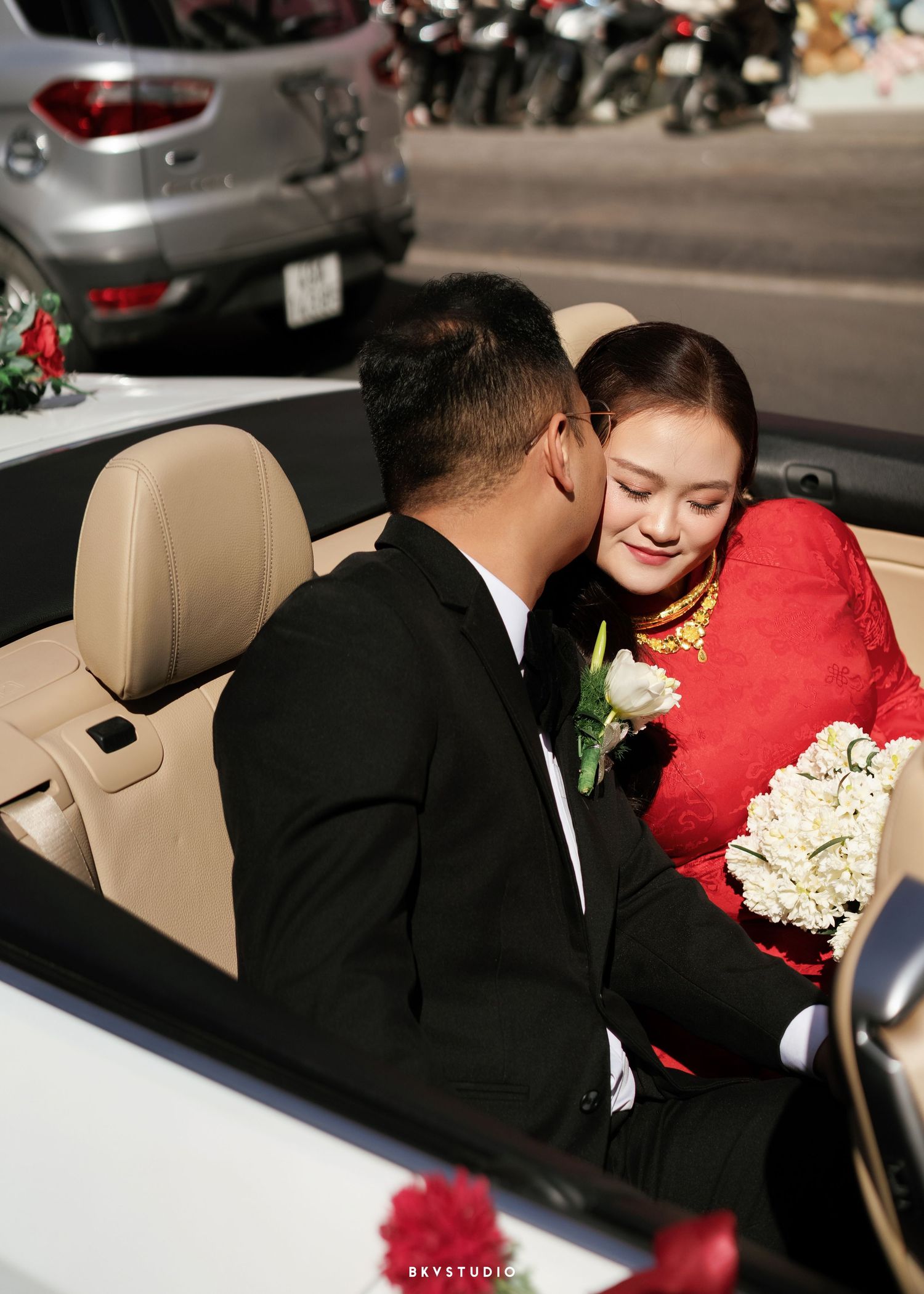 Bride in red ao dai holding white roses receives a kiss from groom in black suit in a white convertible car.