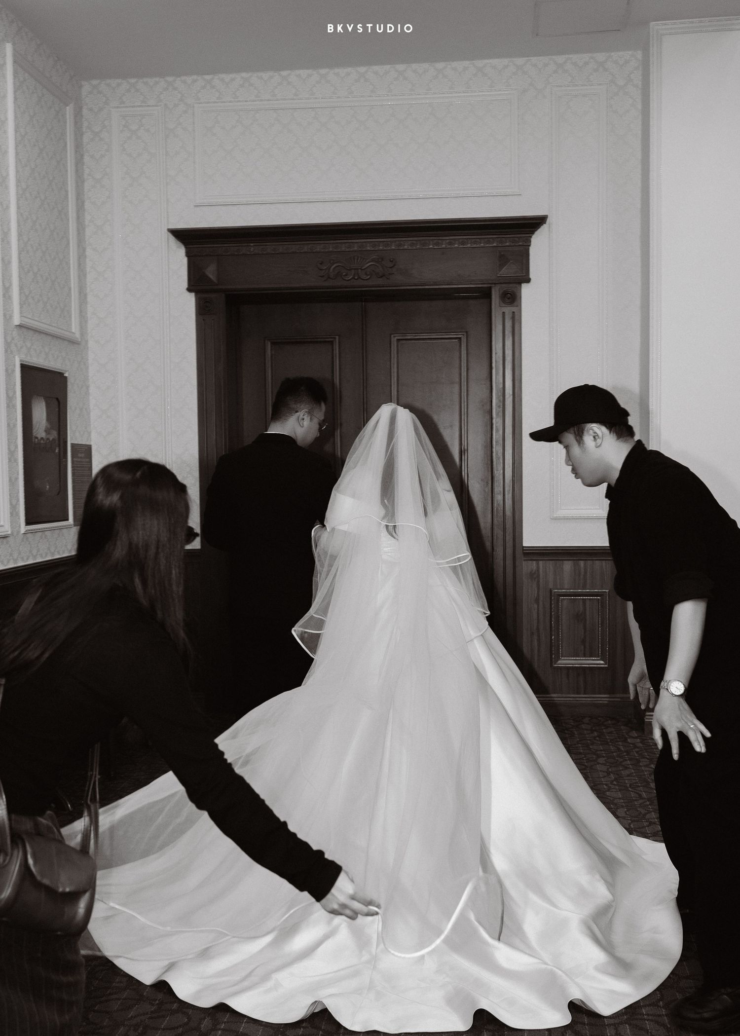 Black and white photo of a bride in a white gown and veil being assisted by attendants before her wedding ceremony.