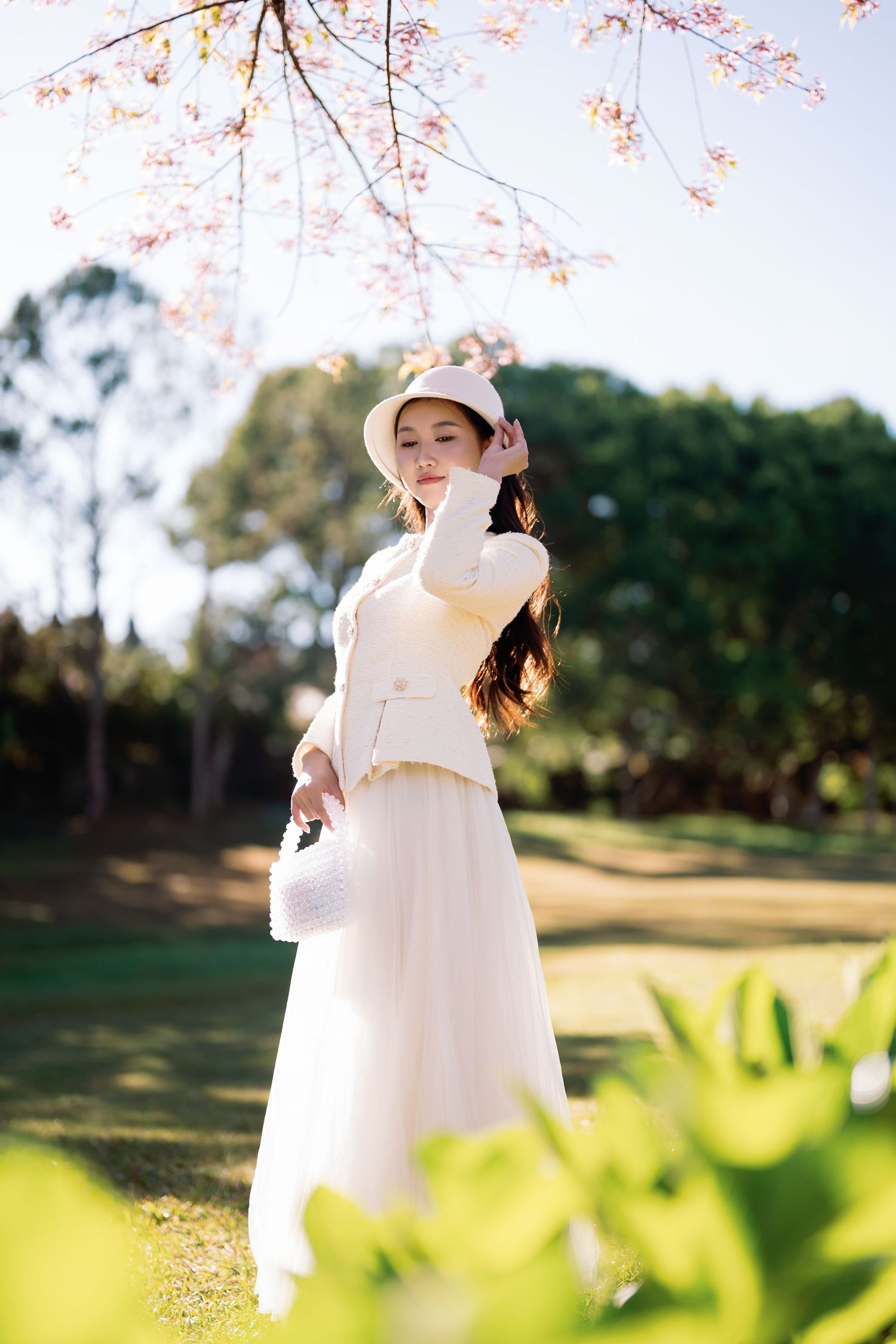 Young woman in white dress and hat poses gracefully under blooming cherry blossom tree in sunny park.
